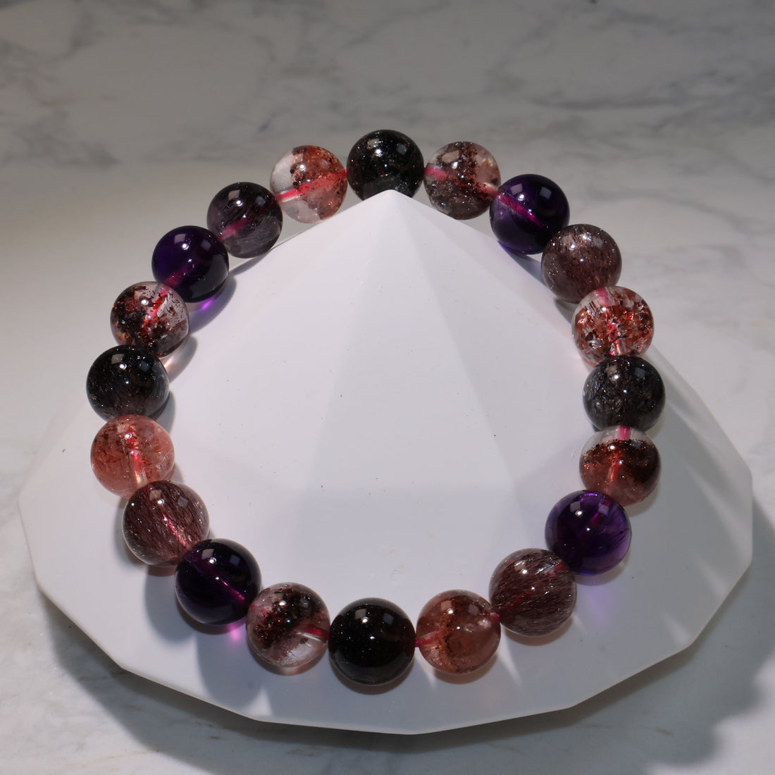 Super Seven bracelet displayed on a marble surface, showing clear, purple, and reddish tones with fine rutile inclusions.