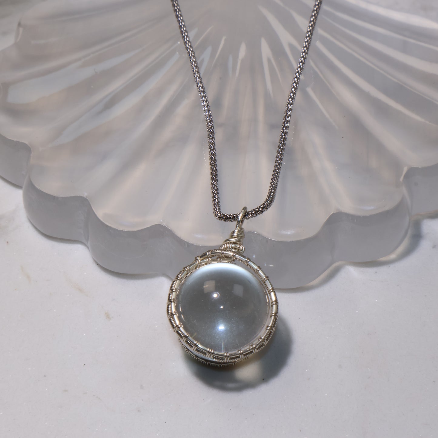 Close-up of the Clear Quartz pendant lying flat on the shell stand, showing its round cabochon shape and polished finish.