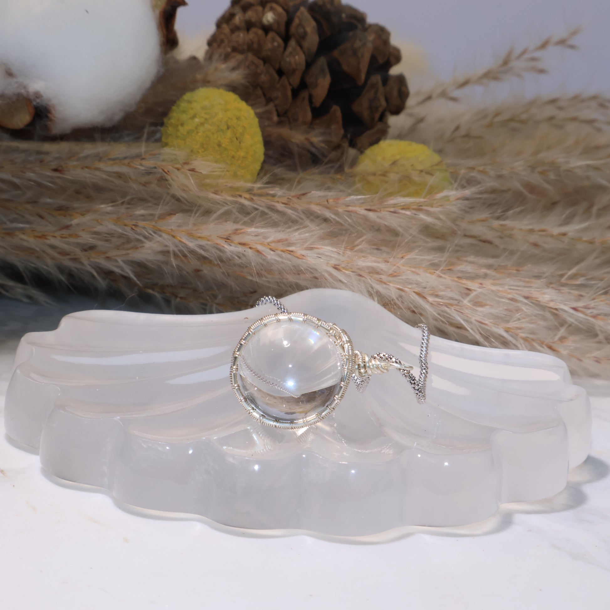 Side angle of the Clear Quartz pendant showing its glass-like transparency and round sphere shape, resting on the shell stand.