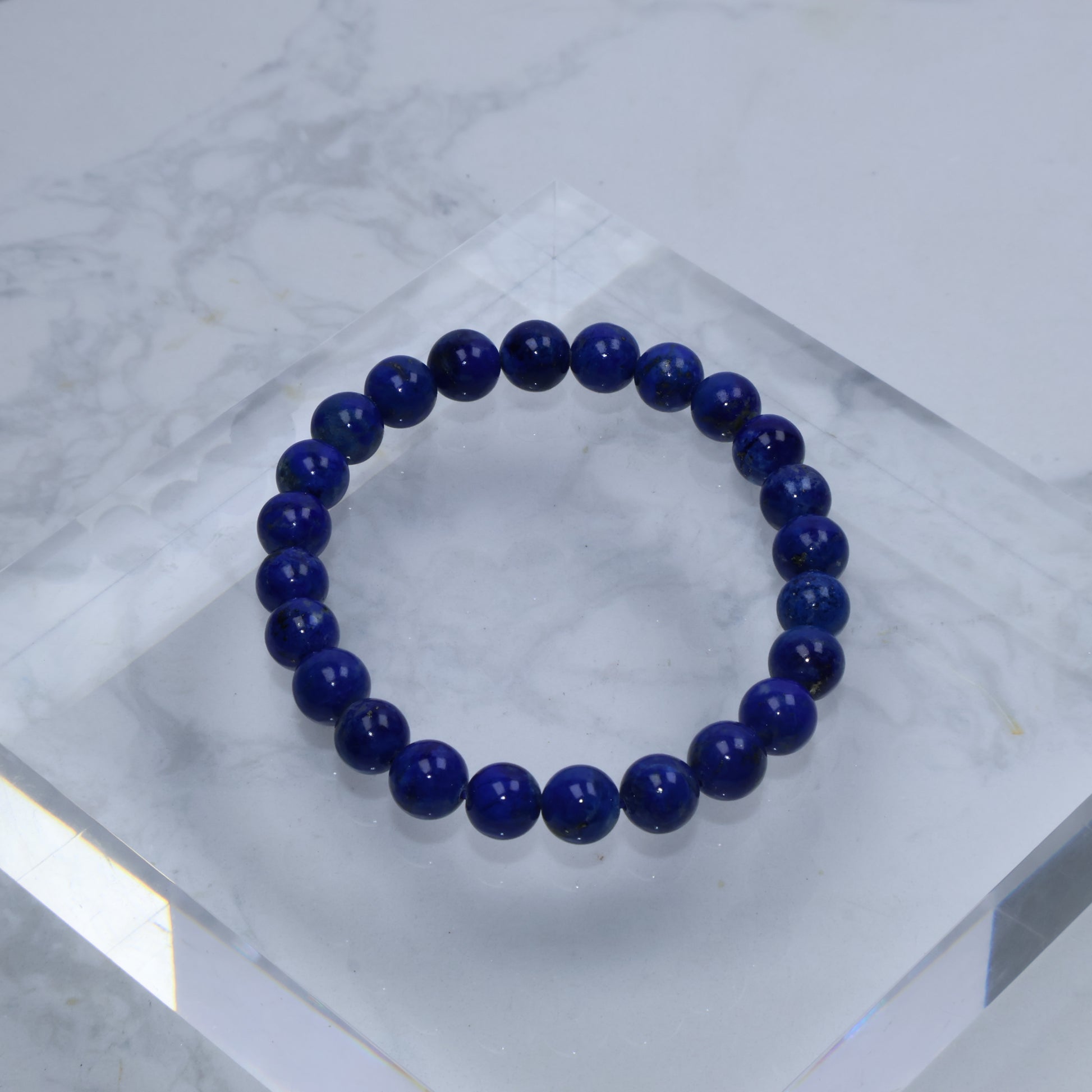 Flat lay of 7mm Lapis Lazuli bracelet on clear acrylic block over white marble background – minimalist product display