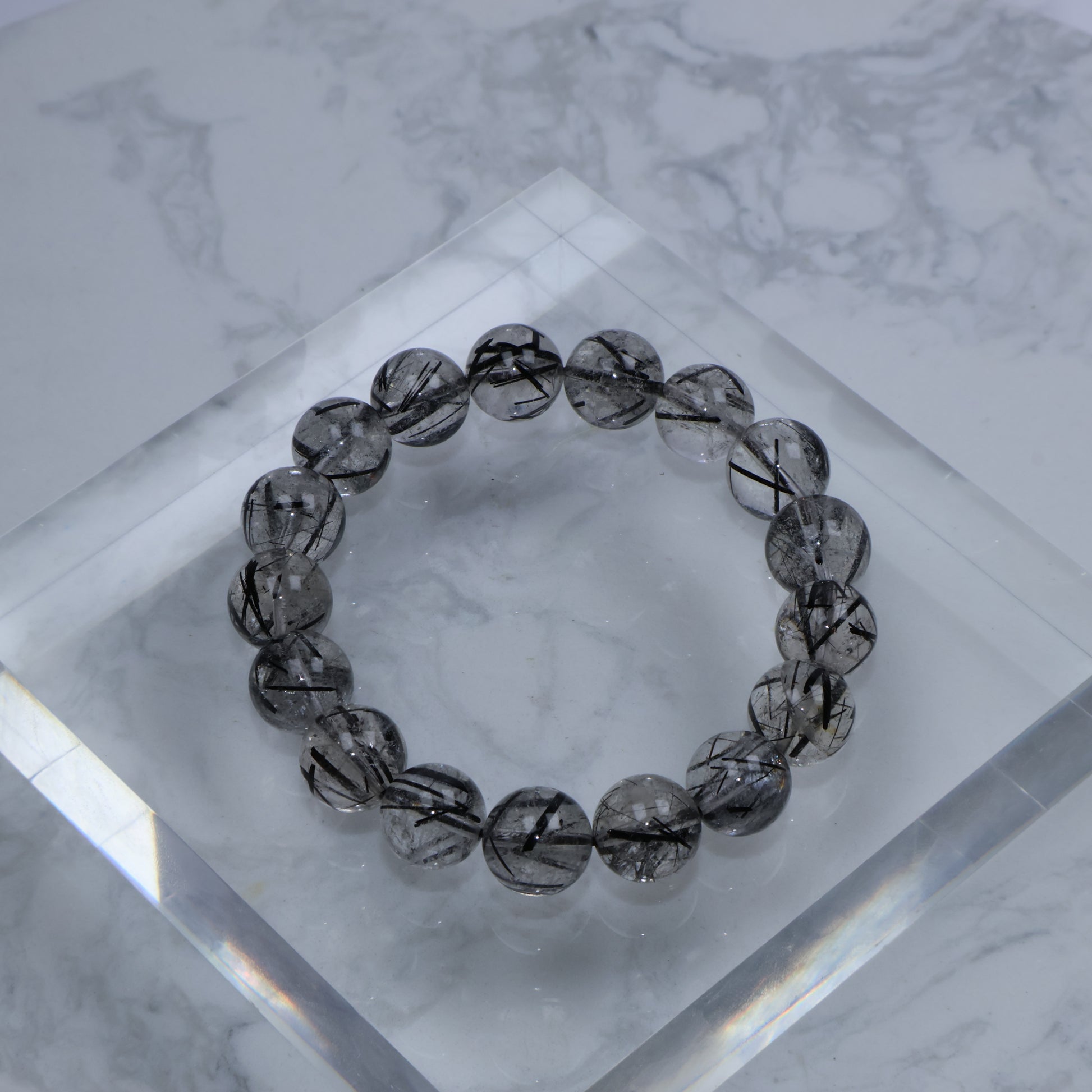 Top-down photo of Black Rutilated Quartz bracelet with bold rutile threads, placed on a clear acrylic block over marble surface