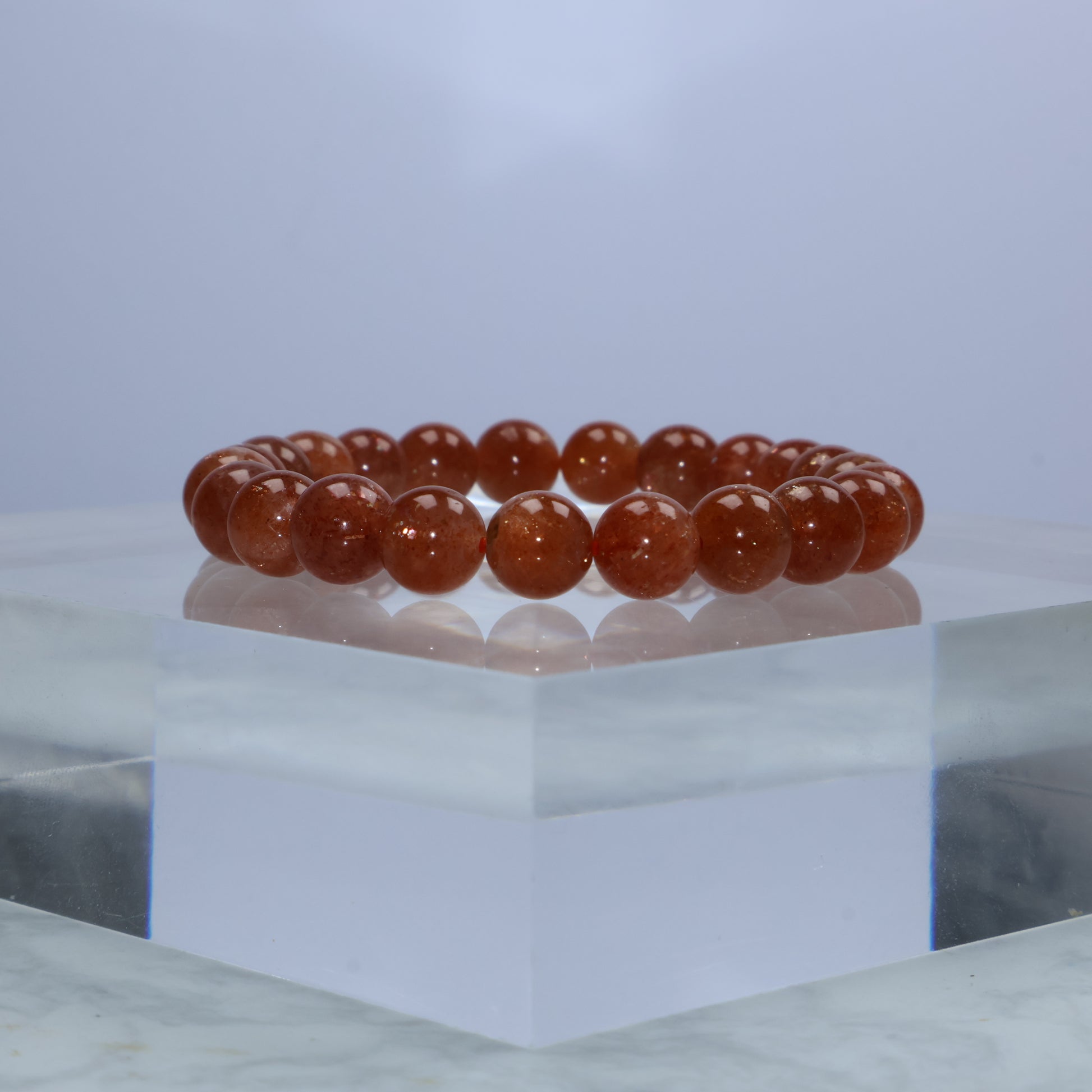 Front-facing image of deep orange 8mm Sunstone bracelet displayed on clear acrylic block, highlighting its vibrant glow.