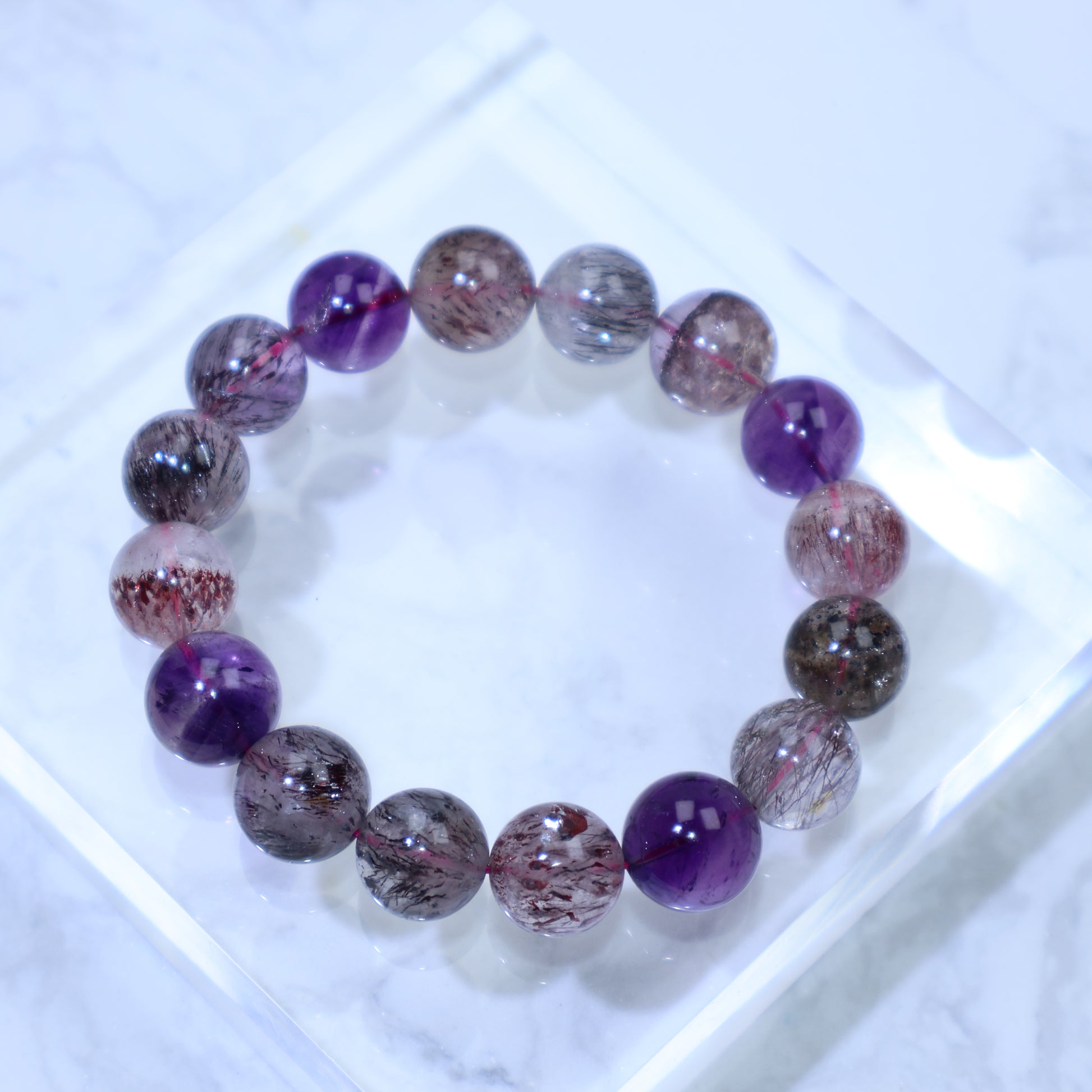 Top-down view of Super Seven (Melody Stone) bracelet on clear acrylic, showing purple quartz and dense inclusion