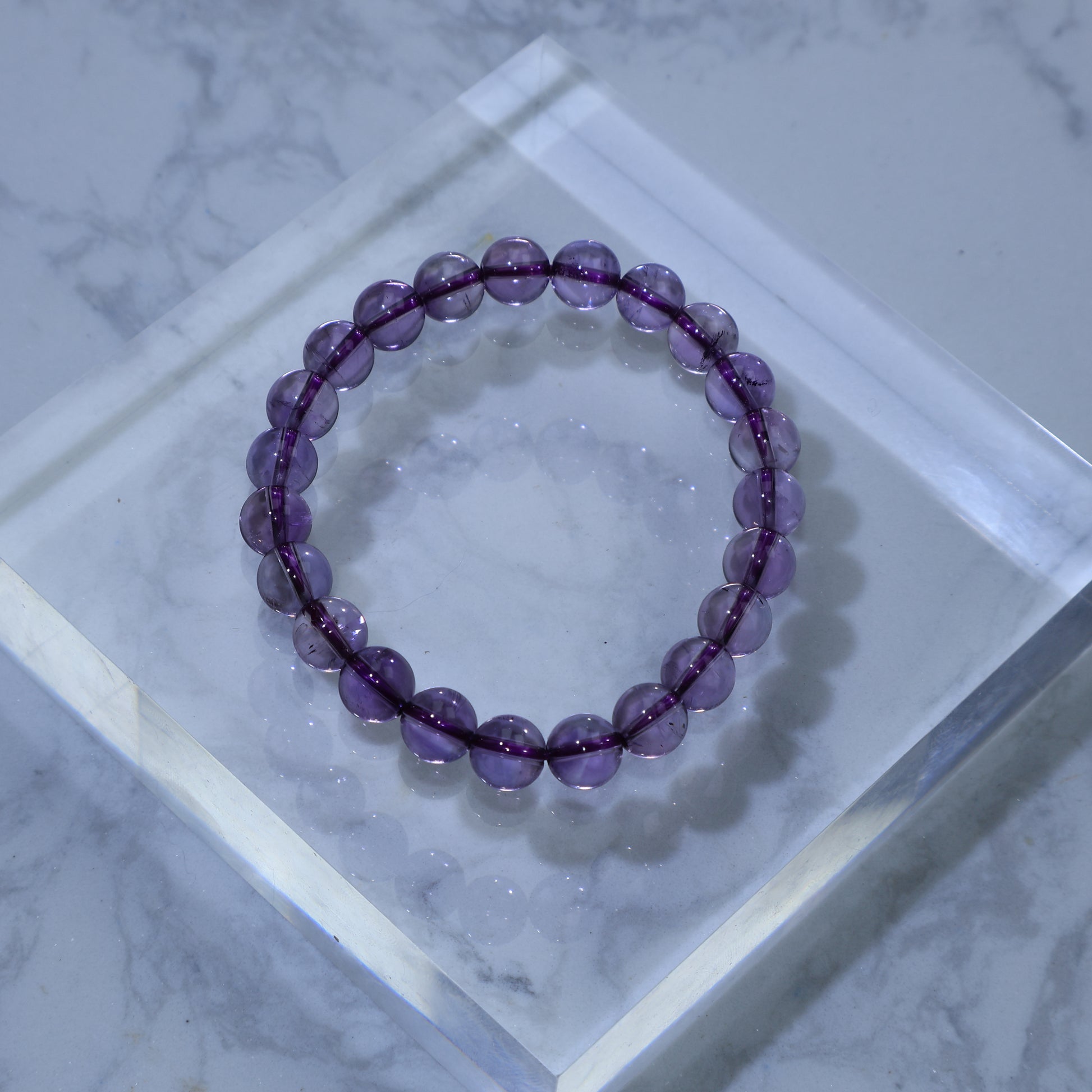 Overhead view of 8mm Amethyst bracelet on a clear acrylic block, glowing with soft violet light