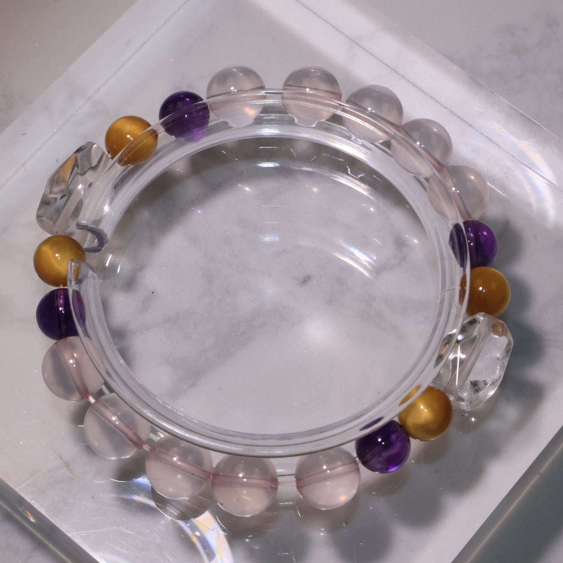 A crystal bracelet with Clear Quartz centerpiece, Rose Quartz, Amethyst, and Tiger’s Eye displayed on a clear stand.