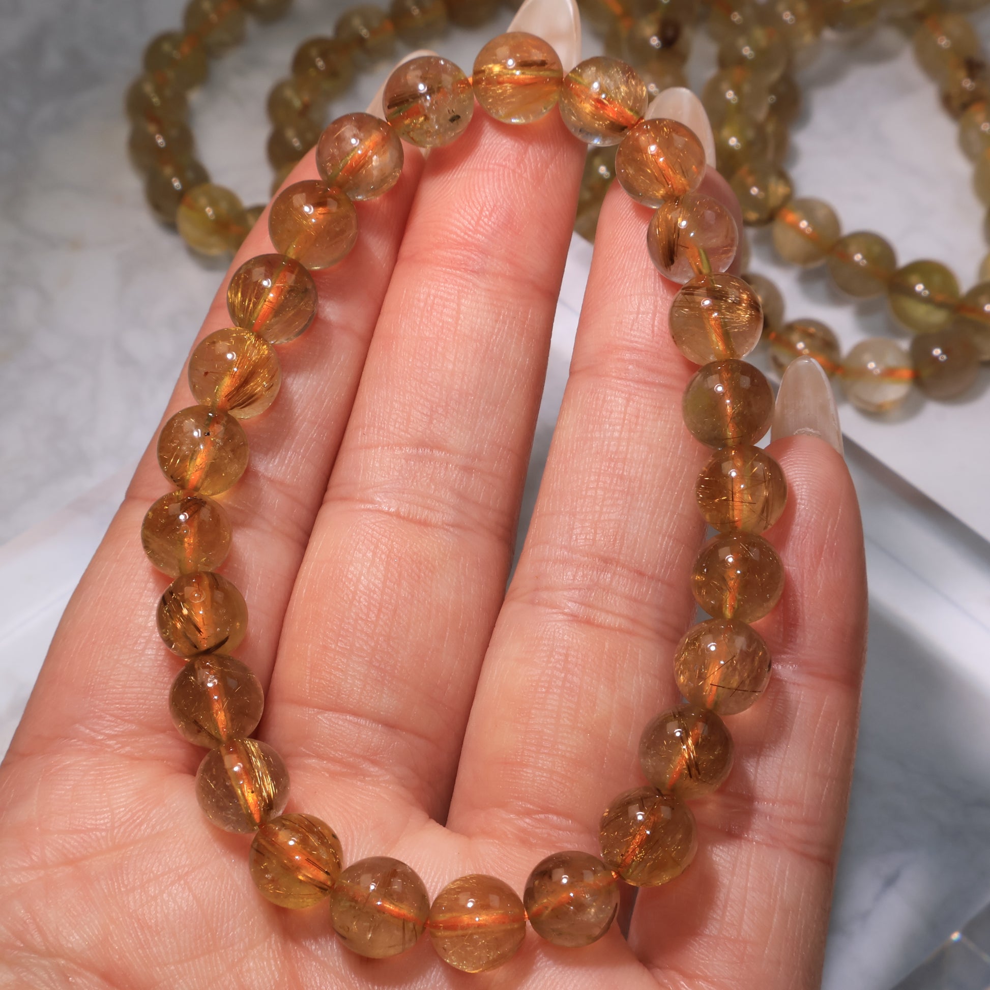 Hand holding a Rutilated Quartz bracelet, showing clear beads with golden and red rutile inclusions.
