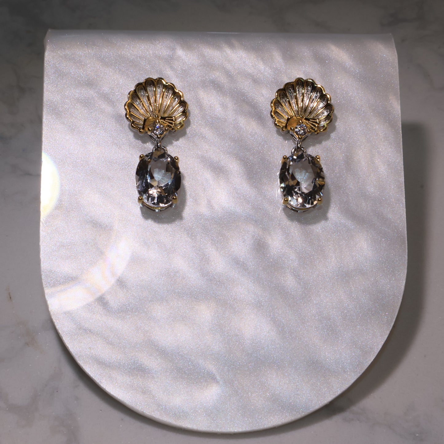 A pair of gold shell-shaped earrings with clear quartz drops displayed on a pearly background.