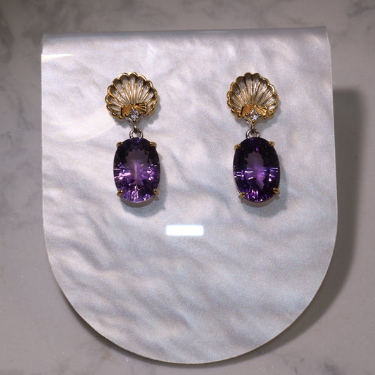 A pair of amethyst drop earrings with golden shell tops and oval purple gemstones displayed on a pearly background.
