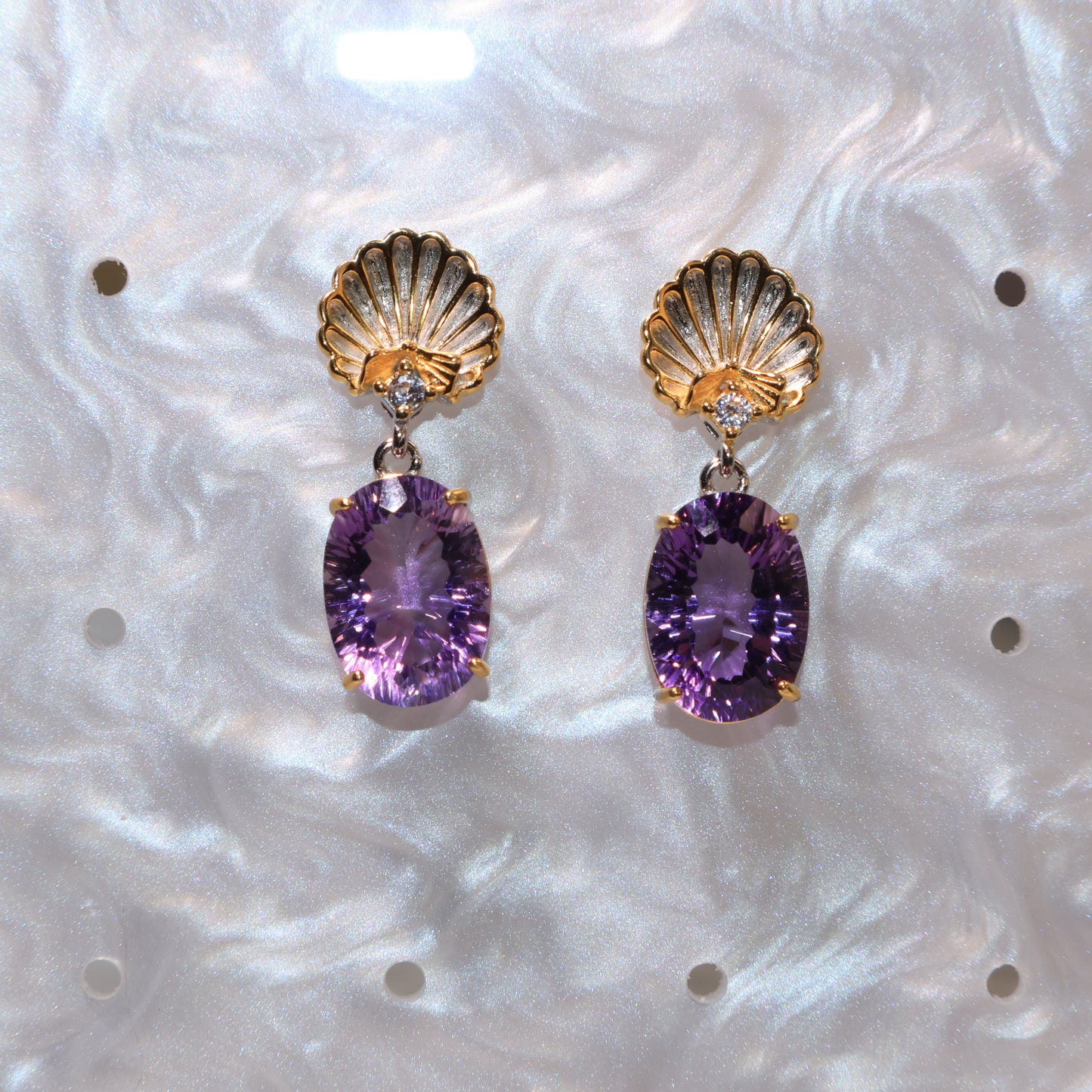 The earrings displayed on a textured white board, emphasizing the contrast between the gold shell and deep purple gem.
