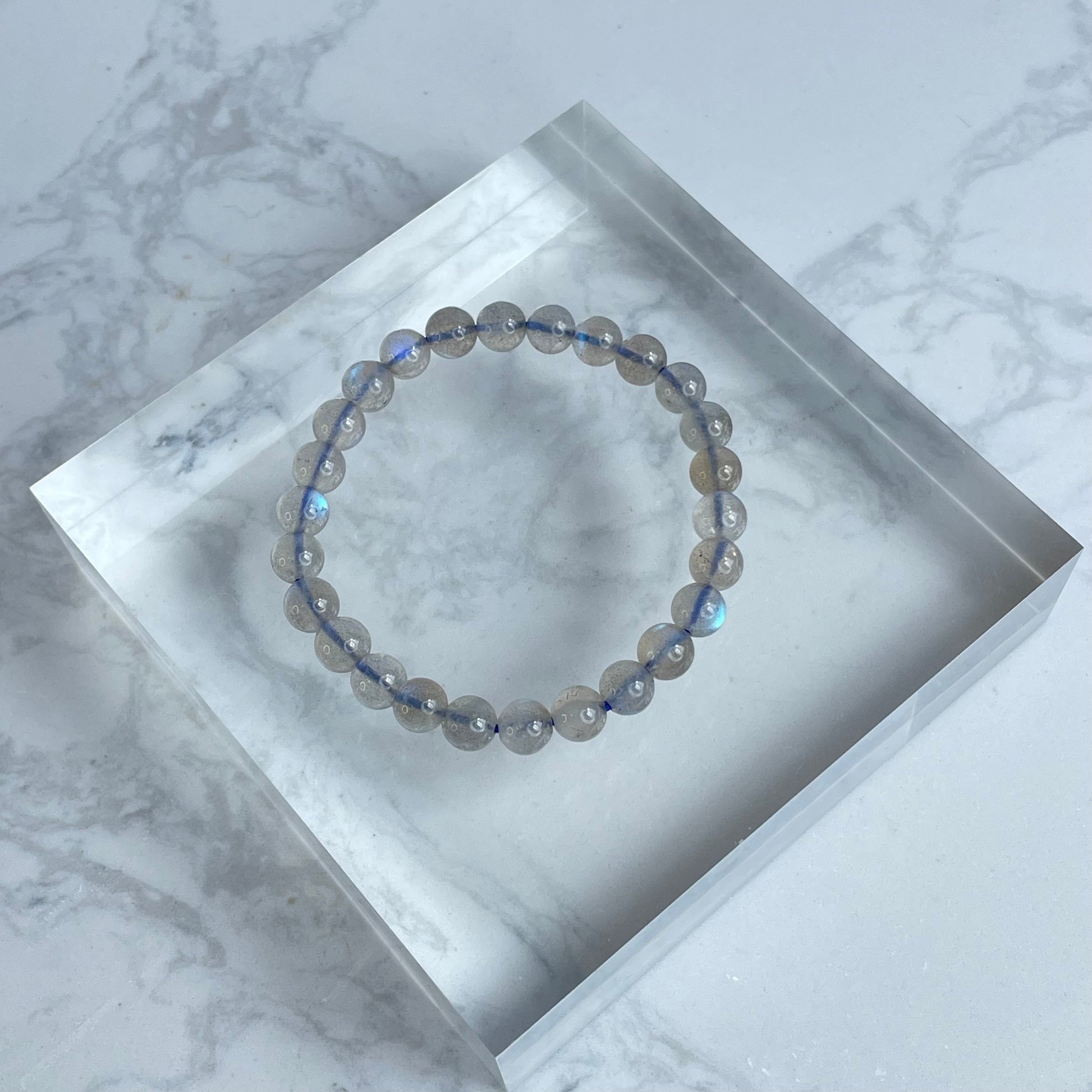 Labradorite bracelet with soft blue glow, displayed on clear acrylic block over marble surface.