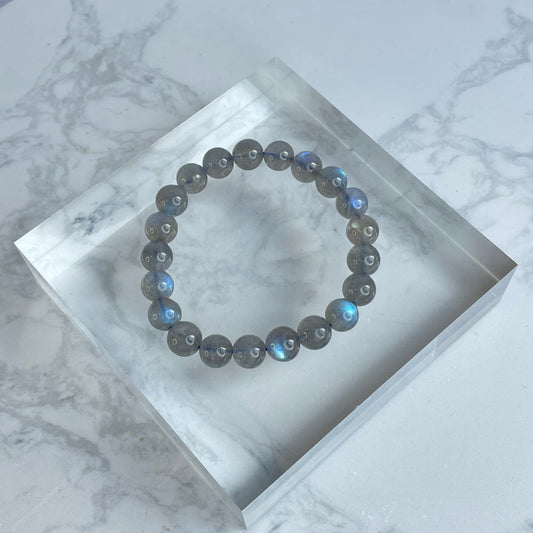 Flat lay of a labradorite bracelet on a clear acrylic display block, featuring round 8mm beads with dark gray tones and striking flashes of blue labradorescence under natural light.