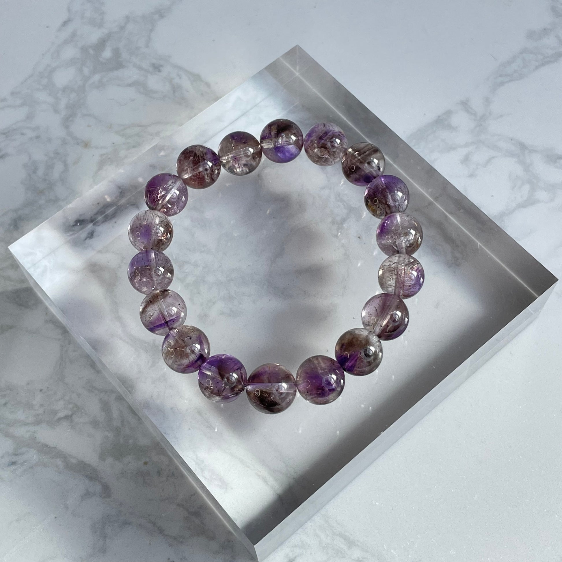 Overhead shot of a round-bead purple Super Seven bracelet placed on a clear acrylic display block, showing a mix of deep purple and reddish mineral inclusions within the transparent beads.
