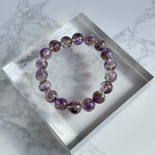 Overhead shot of a round-bead purple Super Seven bracelet placed on a clear acrylic display block, showing a mix of deep purple and reddish mineral inclusions within the transparent beads.