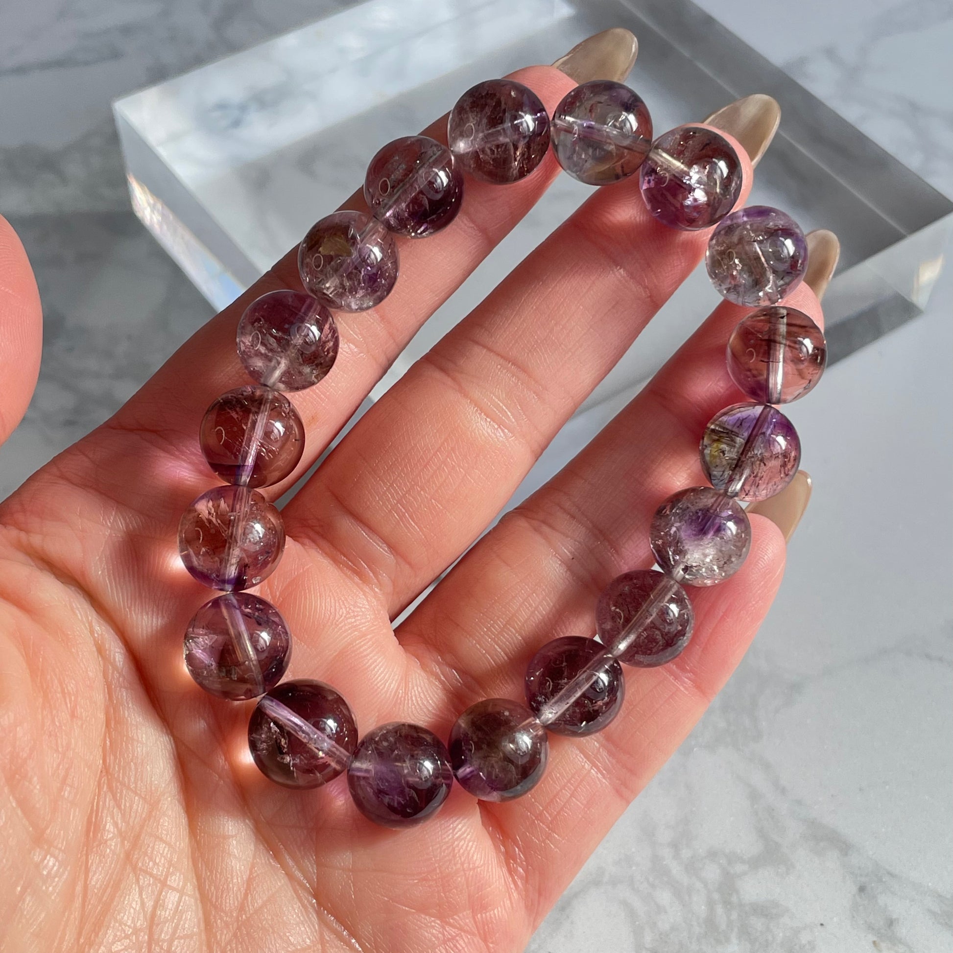 A hand holding a round-bead purple Super Seven bracelet under natural light. The beads are semi-transparent with visible dark purple, red, and silvery mineral inclusions.