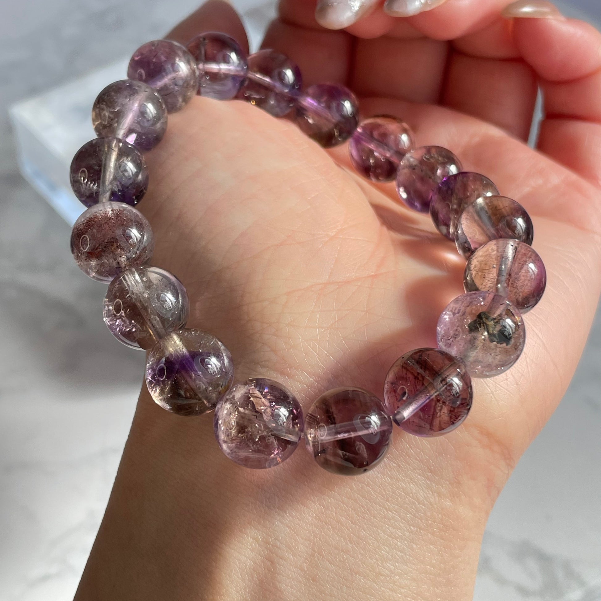 A close-up of a hand wearing a round-bead purple Super Seven bracelet. The beads are translucent with visible mineral inclusions in shades of purple, red, and brown, reflecting under sunlight.