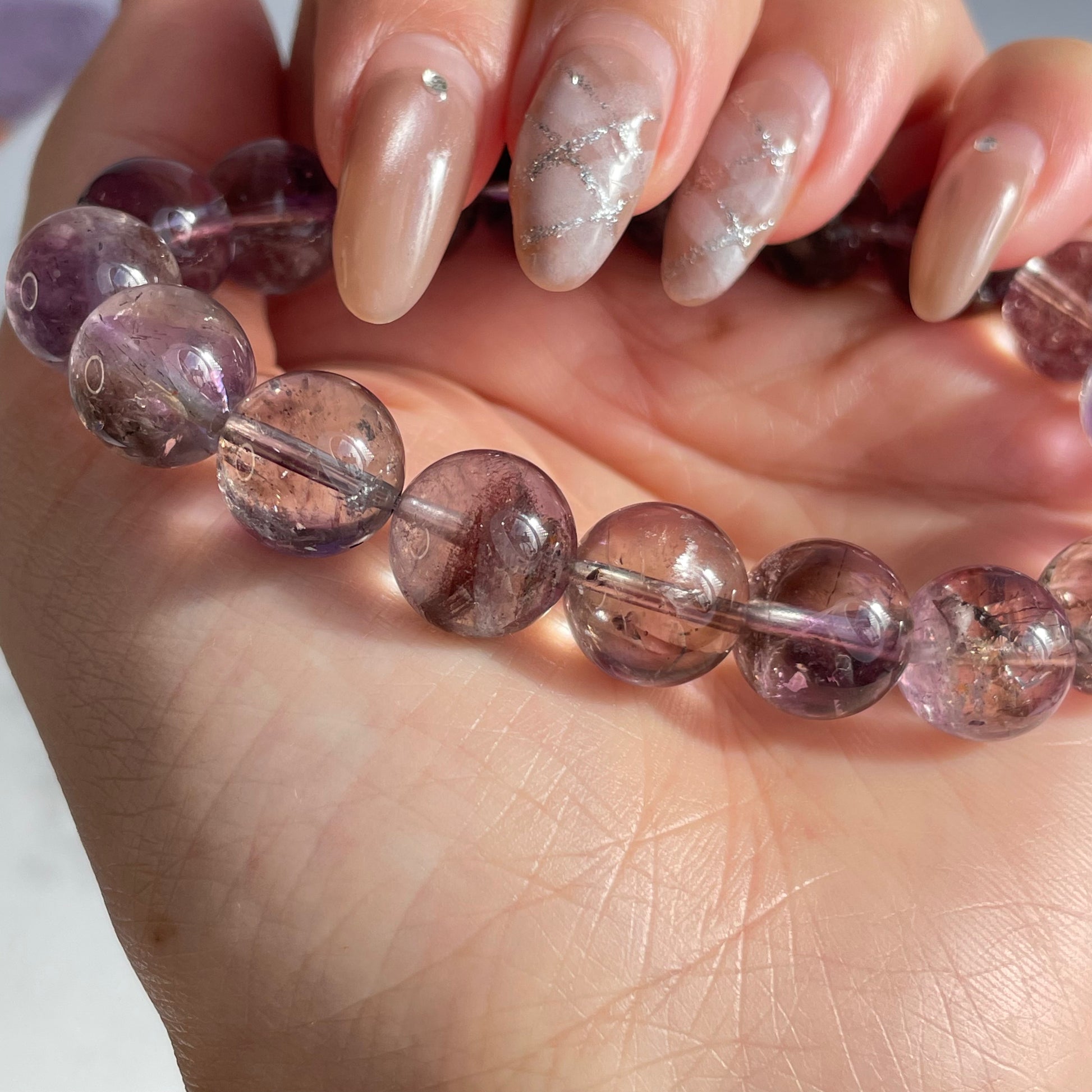 Close-up of a hand holding a round-bead Super Seven bracelet. The beads are semi-transparent with internal inclusions in reddish-purple tones, catching the light beautifully. Fingernails are manicured with a nude and silver design