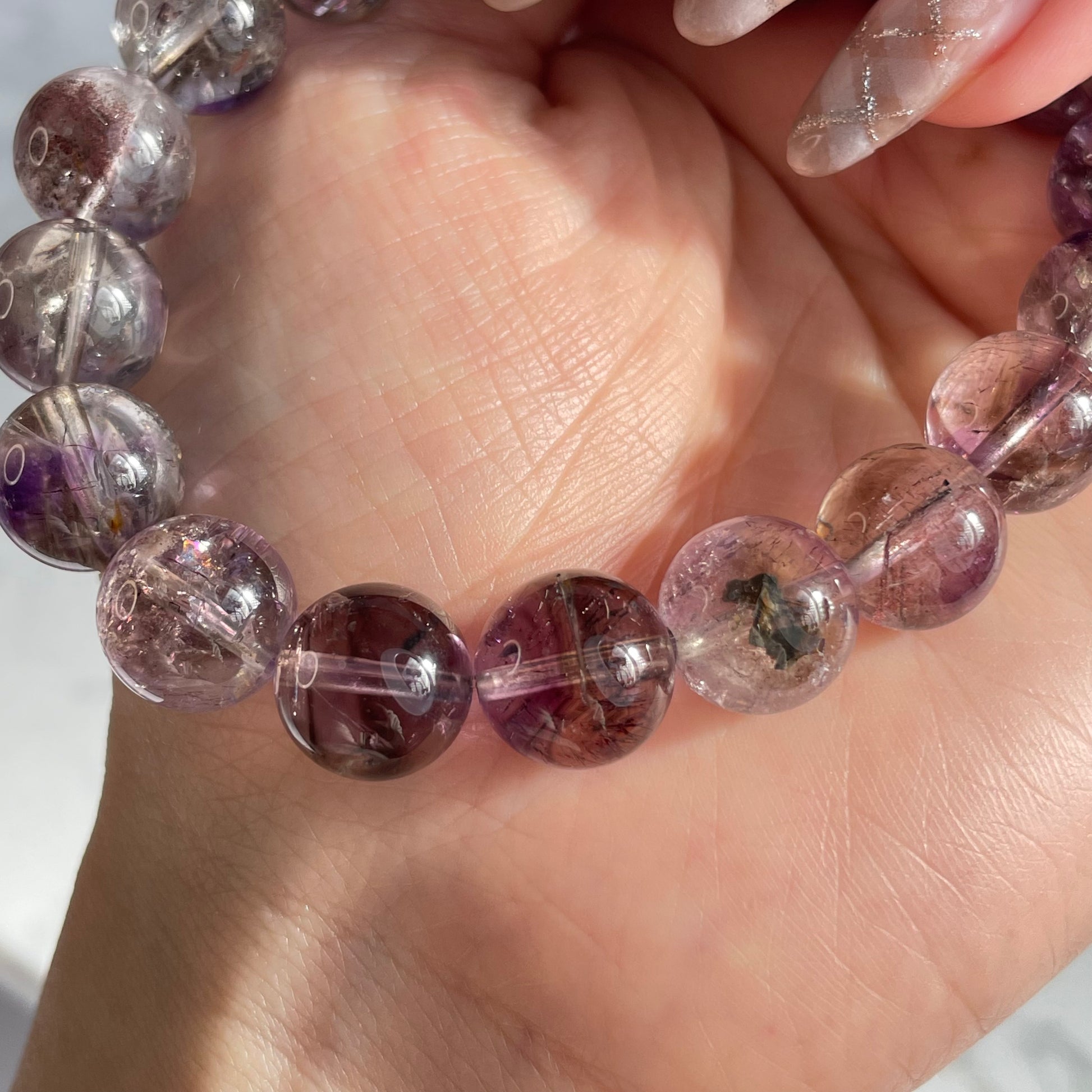 Close-up of a hand holding a round-bead Super Seven bracelet. The beads are mostly transparent with rich inclusions in purple, red, and black tones. One bead shows a prominent black mineral inclusion. The lighting enhances the clarity and internal structure of the crystals.