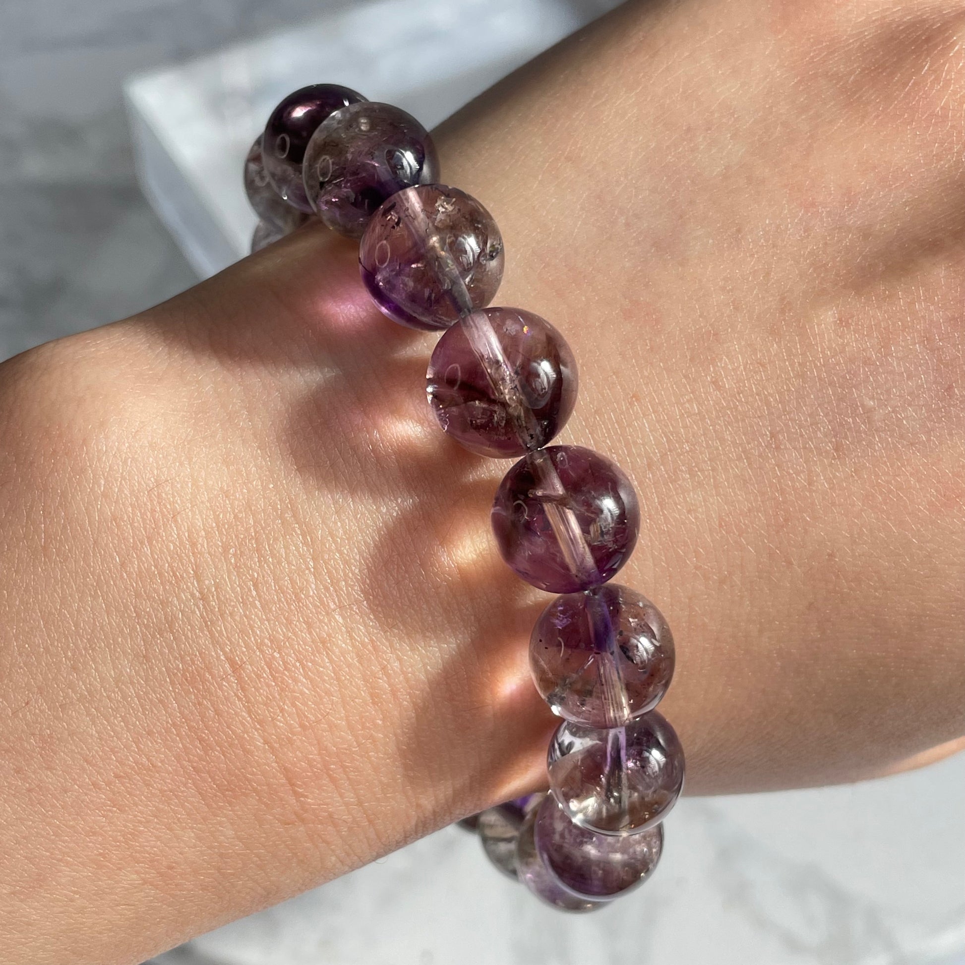Close-up of a round Super Seven crystal bracelet worn on a wrist. The beads are semi-transparent with a mix of deep purple and reddish inclusions, reflecting light and revealing their internal textures.