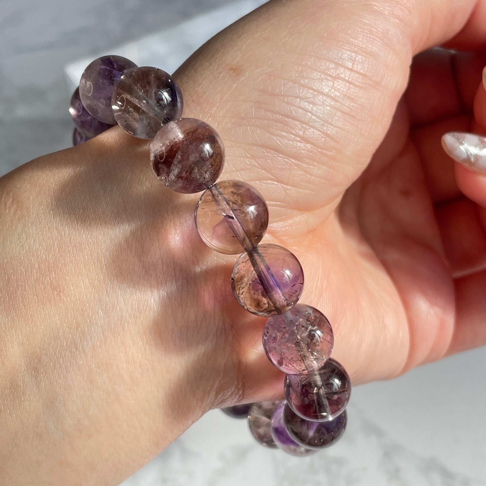 Close-up view of a Super Seven crystal bracelet worn on a wrist, showing semi-transparent round beads with visible internal inclusions in shades of purple, red, and smoky brown under natural lighting.