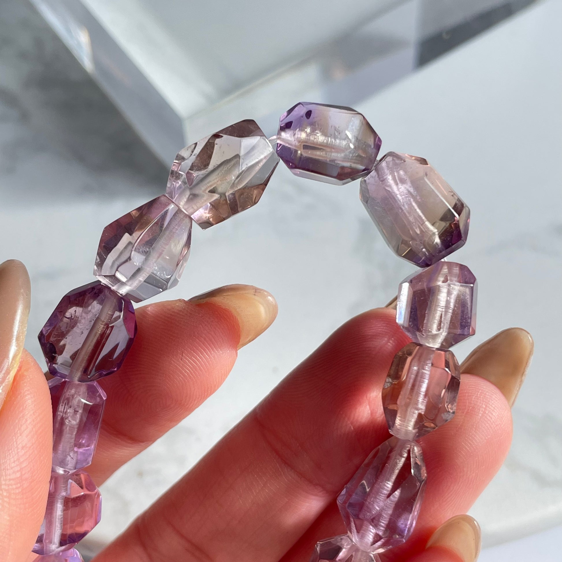 Close-up of a raw-cut ametrine bracelet in hand, showcasing multi-faceted transparent beads with light purple and honey tones under natural sunlight.