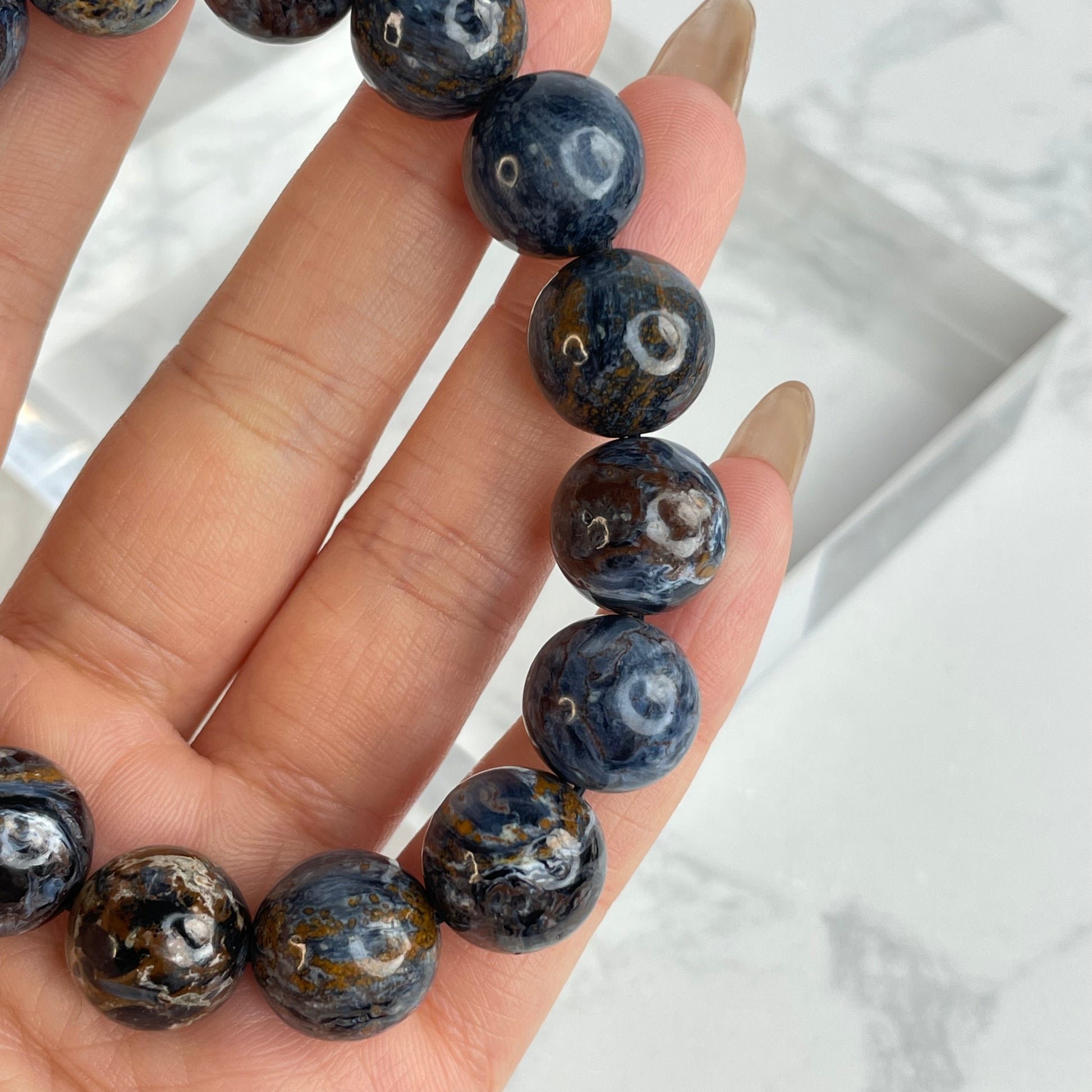Close-up view of 13mm Pietersite bracelet beads, showcasing natural swirling patterns in deep blue, brown, gold, and white tones, held between fingers.
