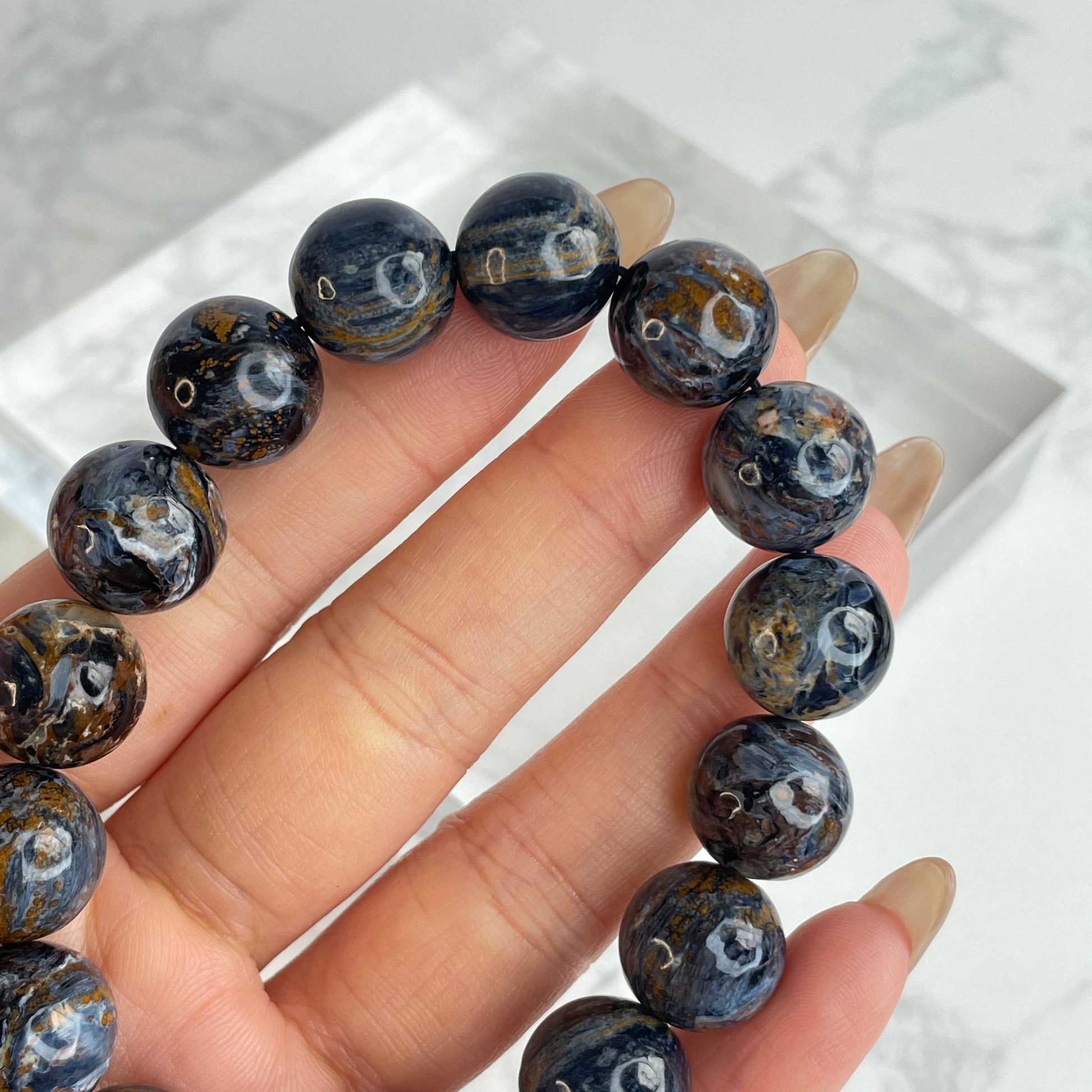 Close-up of 13mm Pietersite bracelet beads held between fingers, highlighting swirling patterns in dark blue, golden brown, and silvery white tones.