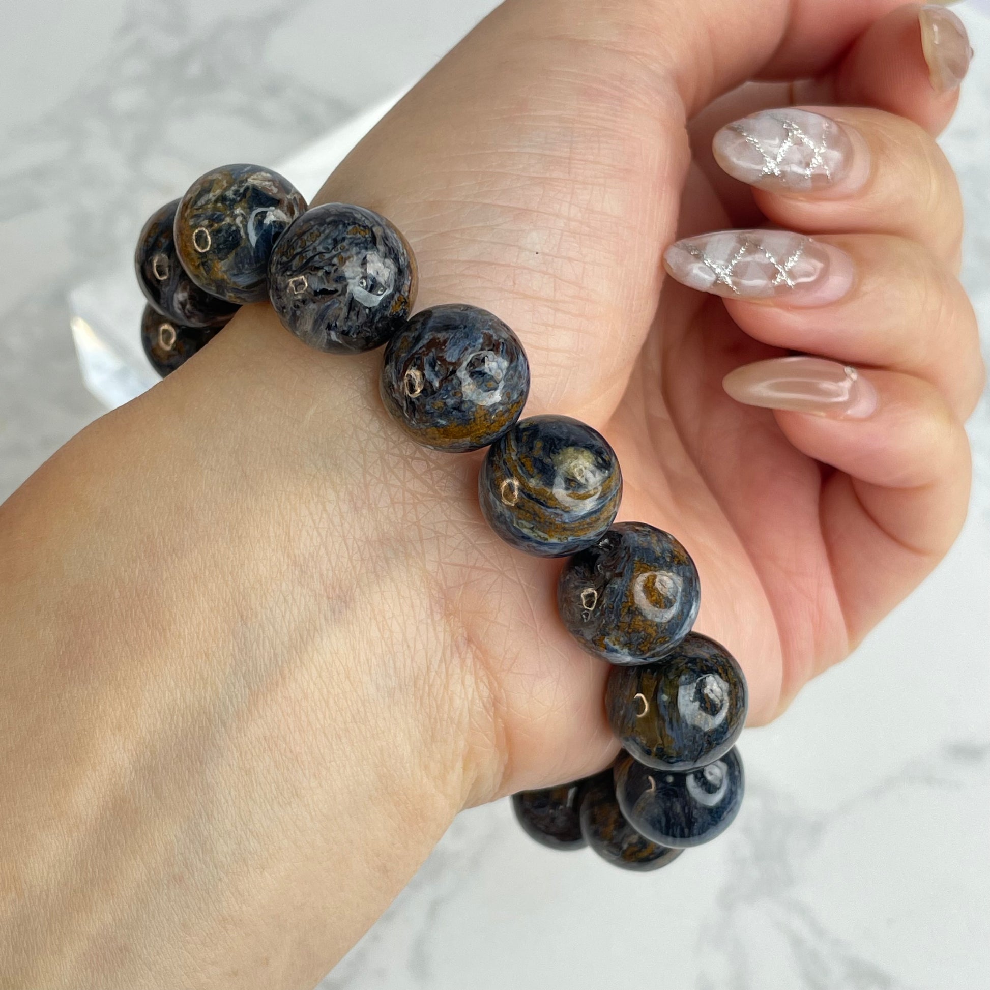 Close-up of a 13mm Pietersite crystal bracelet from Namibia, featuring dark blue and golden brown swirls, worn on a woman’s wrist with manicured nails.