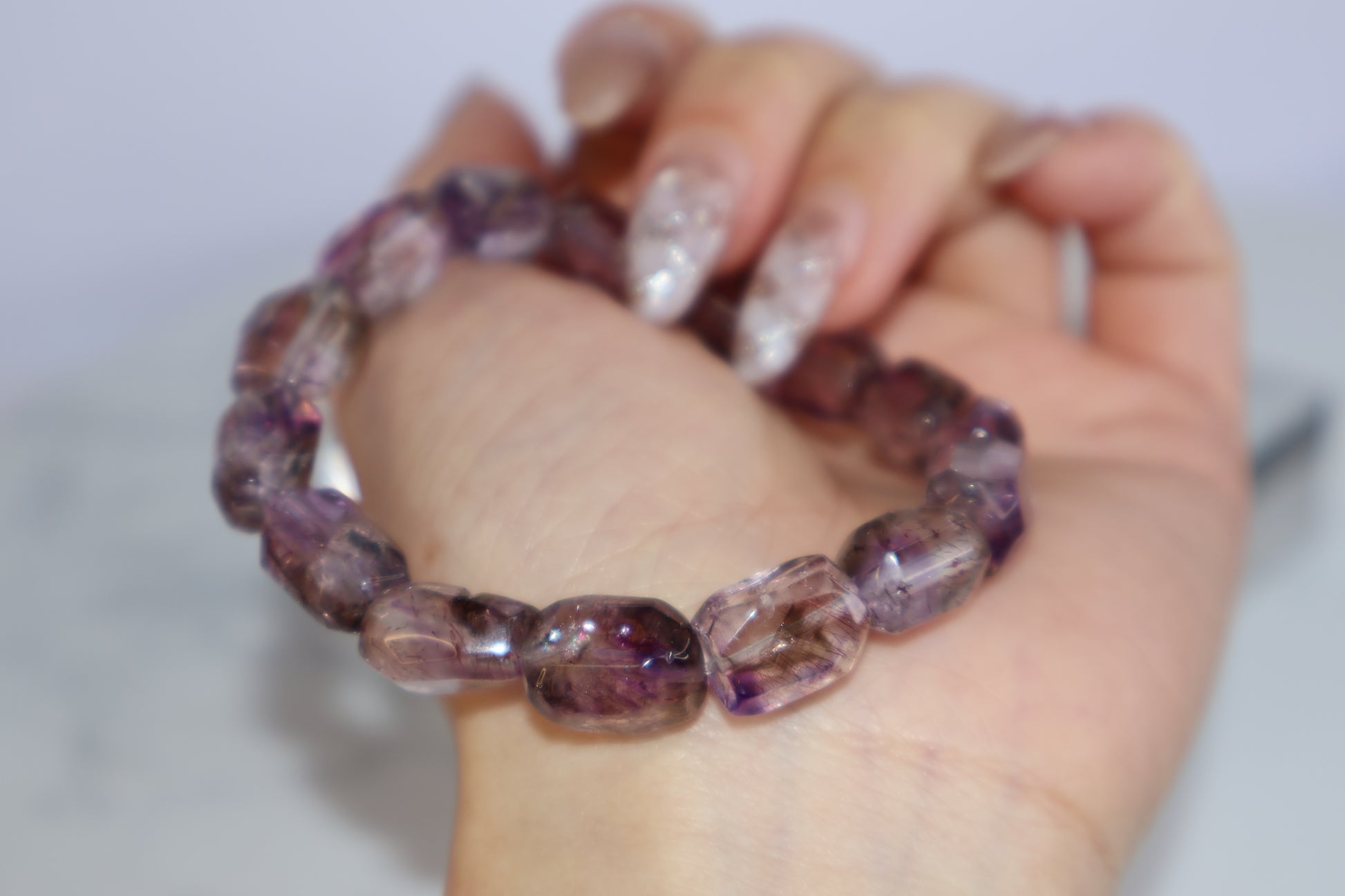Close-up of a hand holding a Super Seven freeform bracelet, with visible rutile strands and shimmering purple hues under natural lighting.