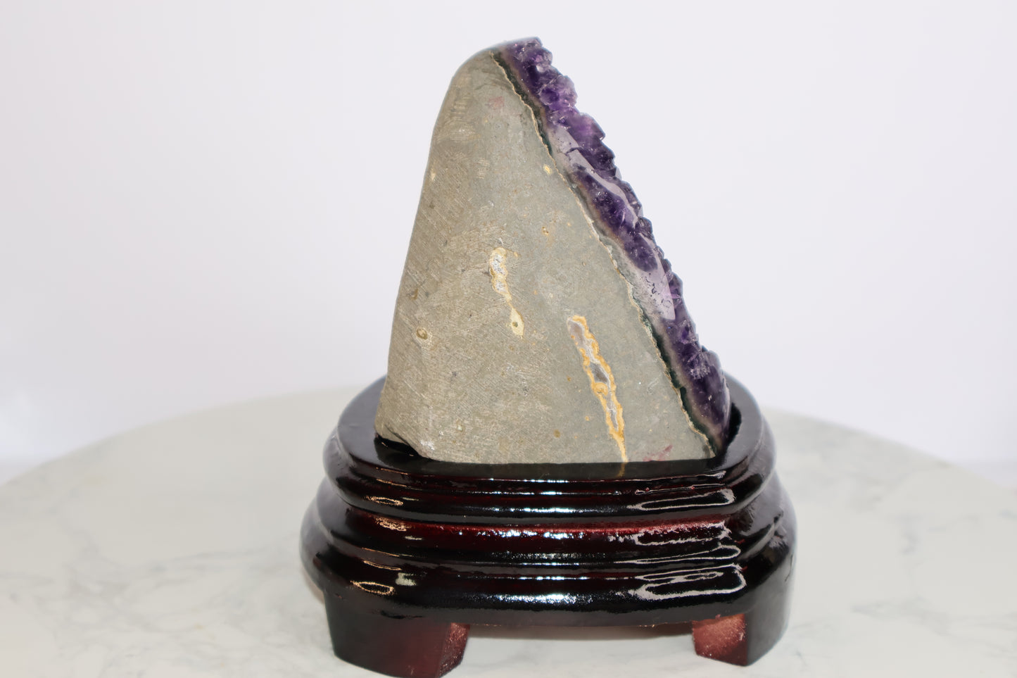 Side view of an amethyst geode showing the raw outer stone surface and the edge of the crystal formation, placed on a glossy dark wood base.