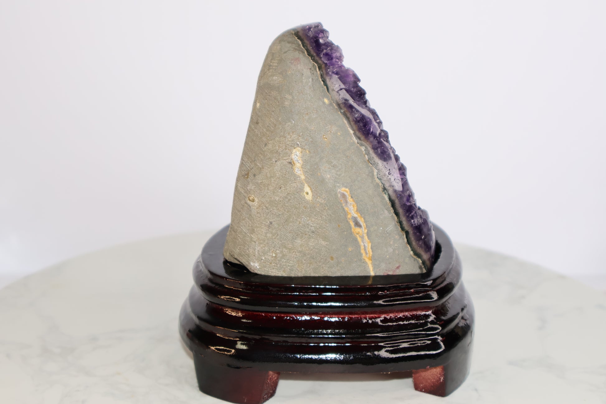 Side view of an amethyst geode showing the raw outer stone surface and the edge of the crystal formation, placed on a glossy dark wood base.