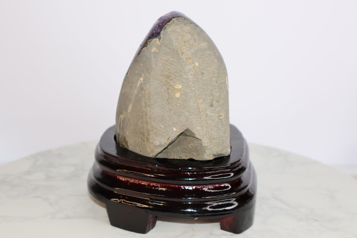Back view of an amethyst geode showing the raw stone surface with natural textures and minor cracks, placed on a glossy dark wood stand.
