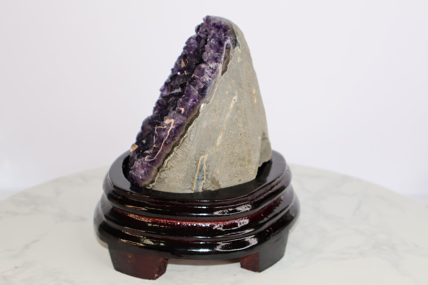 Right side view of an amethyst geode showing the rough grey stone with streaks and the edge of the purple crystal cluster, set on a glossy wooden base.
