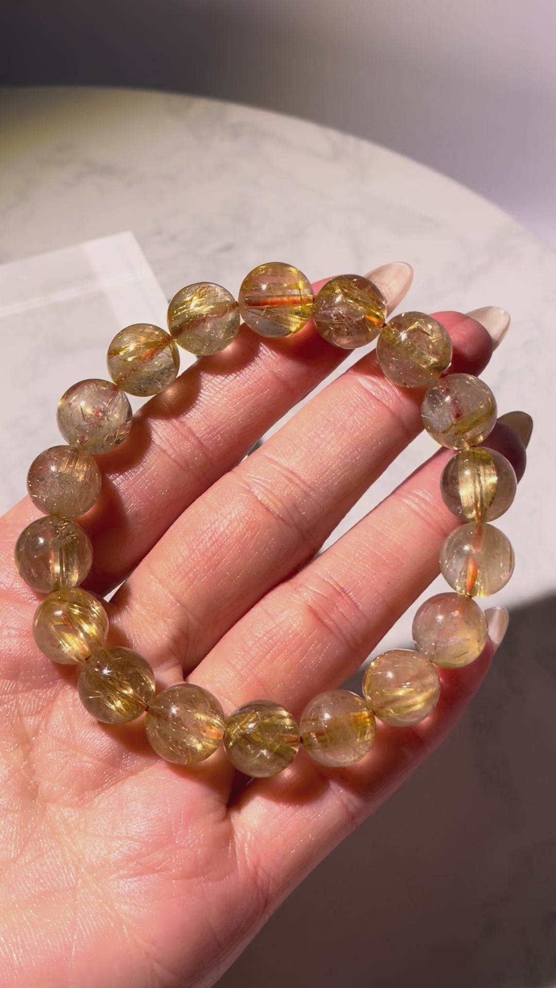 Video of an 11mm Golden Rutilated Quartz bracelet with red hair-like inclusions, rotating under natural light. Each bead sparkles with golden clarity and distinctive rutile threads, highlighting the rich golden and copper-red tones in motion.