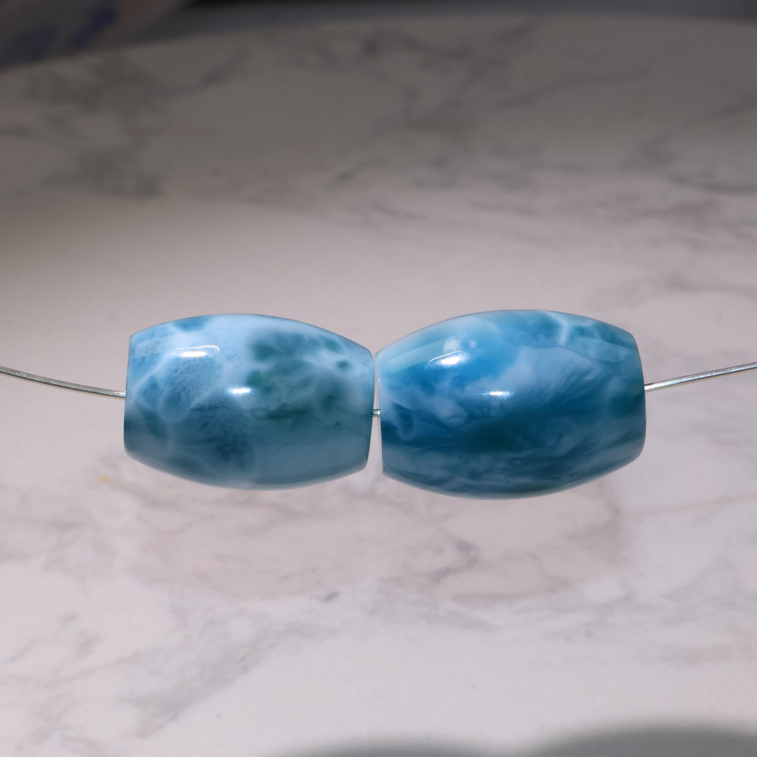 Two polished barrel-shaped Larimar beads in sky blue with white cloud-like patterns, strung on a wire, displayed on a marble background.