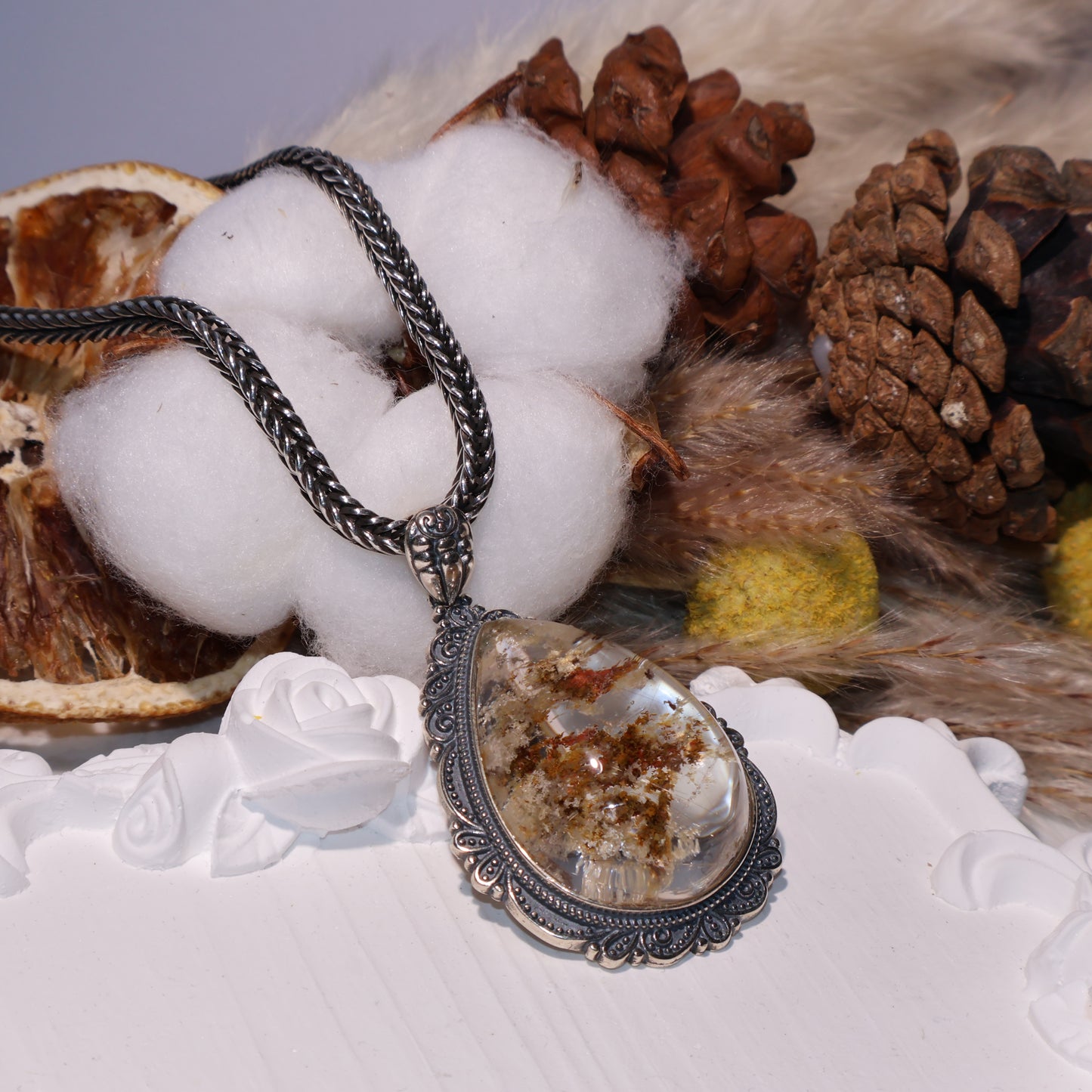 Close-up of teardrop-shaped Phantom Quartz pendant with red and yellow inclusions, set in an ornate vintage S925 silver frame. Dried flowers and pinecones in the background.