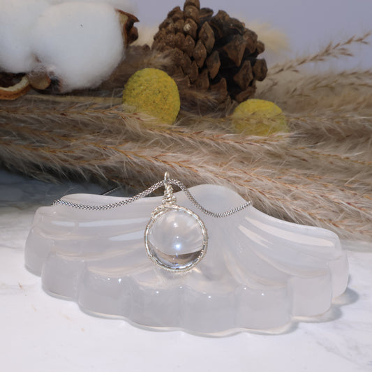 A 17.6mm Clear Quartz pendant with handmade silver-tone wire wrapping, displayed on a translucent shell-shaped stand with natural cotton and pinecone decor in the background.