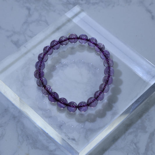 Overhead view of 8mm Amethyst bracelet on a clear acrylic block, glowing with soft violet light