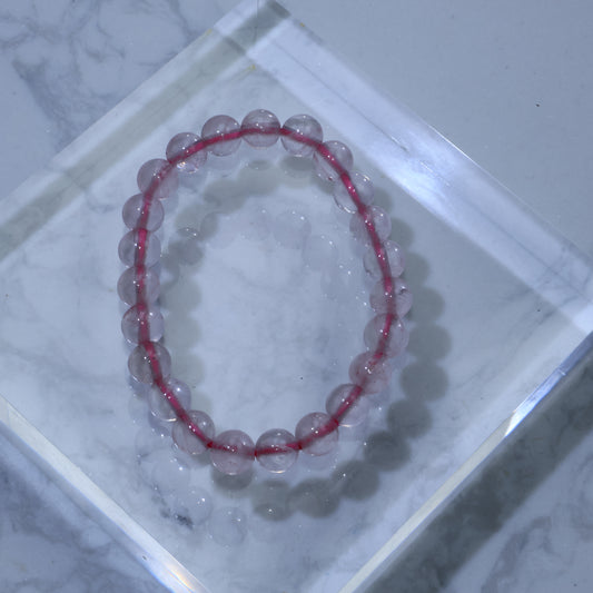Top-down view of Rose Quartz bracelet with 8mm round beads, showing soft pink hue and natural ice cracks, displayed on acrylic block over marble background.