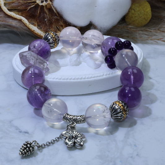 Styled shot of Rose Quartz × Amethyst bracelet on white display; shows double-terminated amethyst point, mini Uruguayan amethyst cluster, and three copper findings with dangling charms.