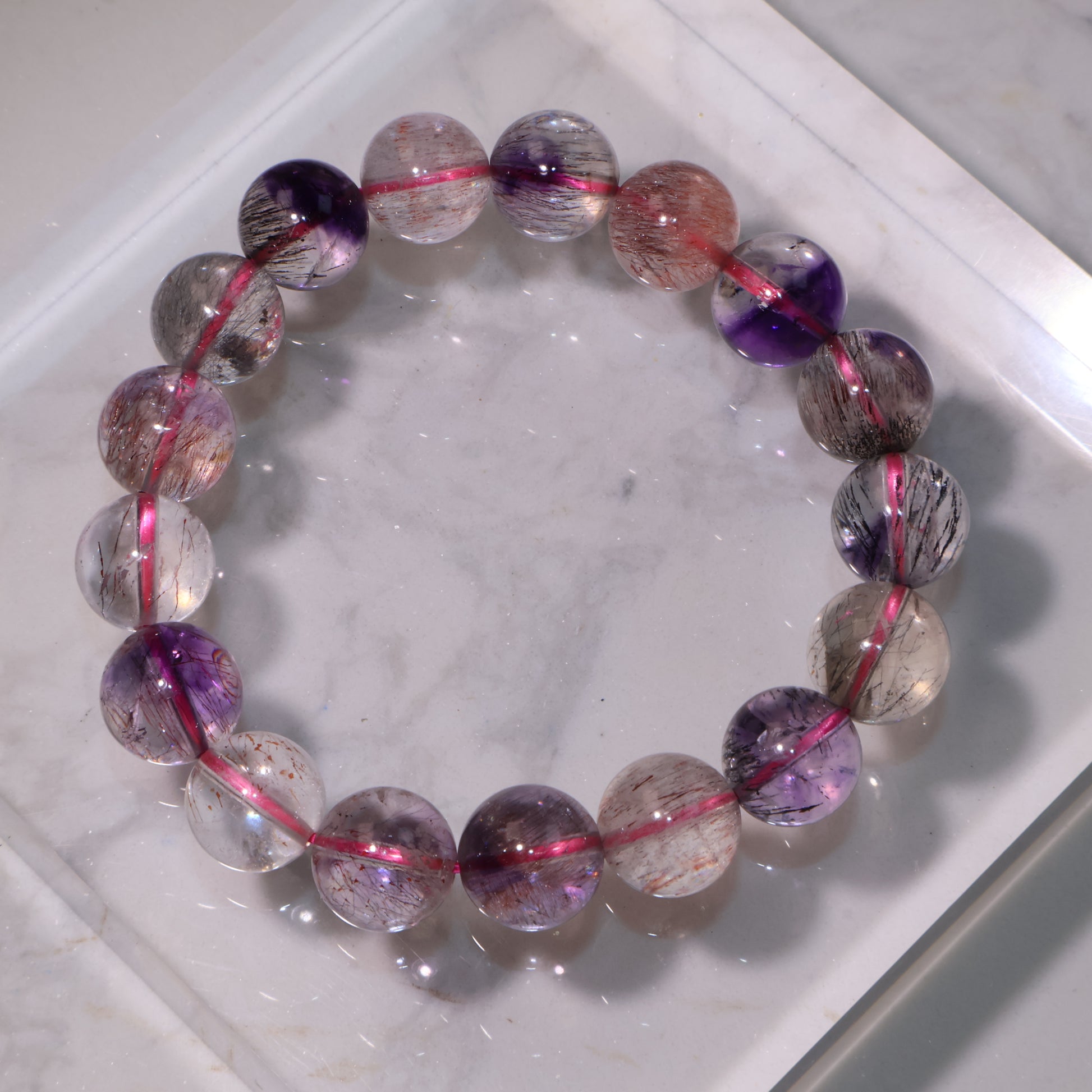 Super Seven bracelet displayed on a marble surface, showing clear, purple, and reddish tones with fine rutile inclusions.