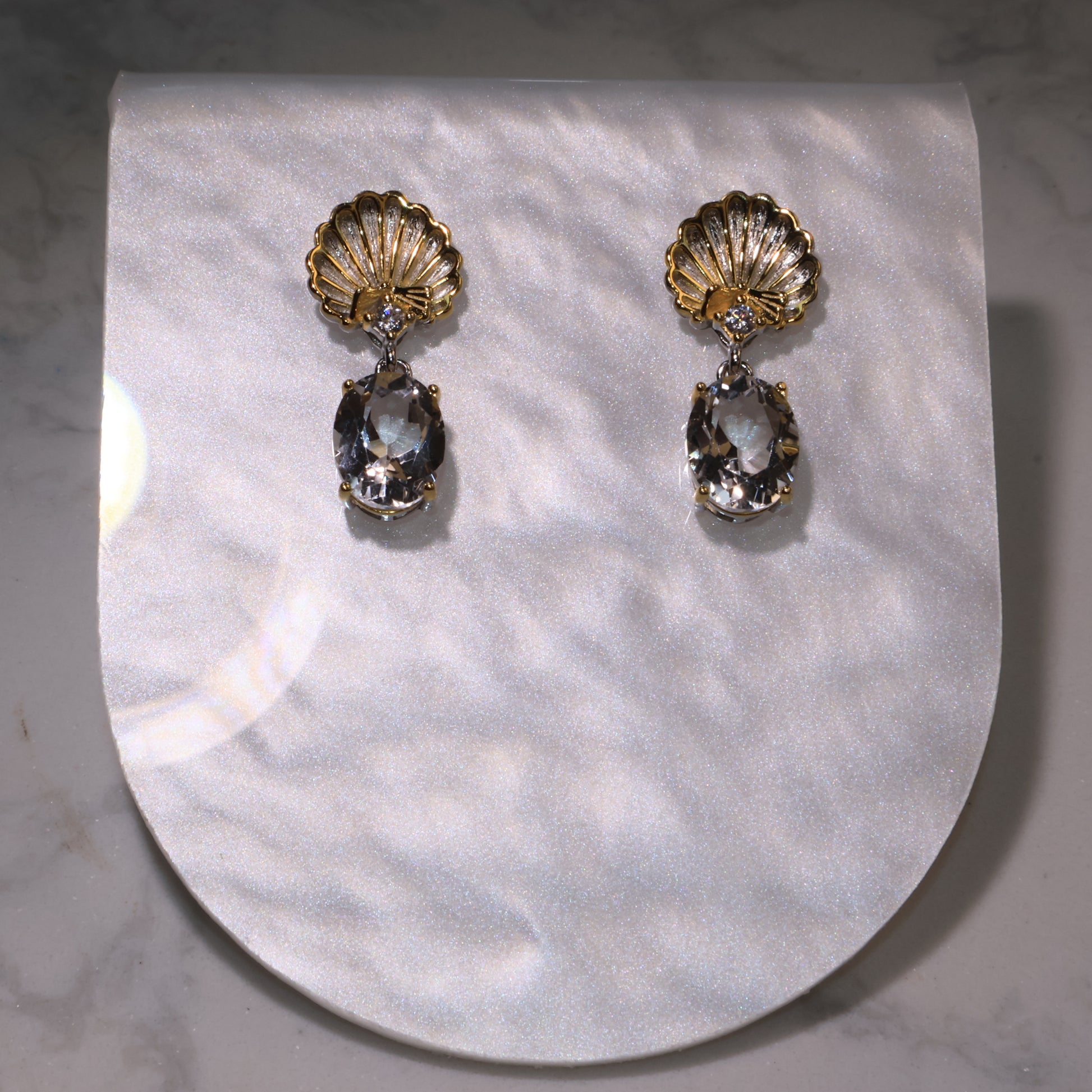 A pair of gold shell-shaped earrings with clear quartz drops displayed on a pearly background.