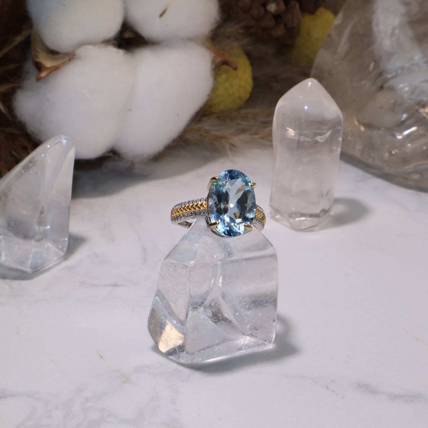 A Blue Topaz ring displayed on a clear quartz base, glowing under soft natural light, surrounded by cotton flowers and crystals.