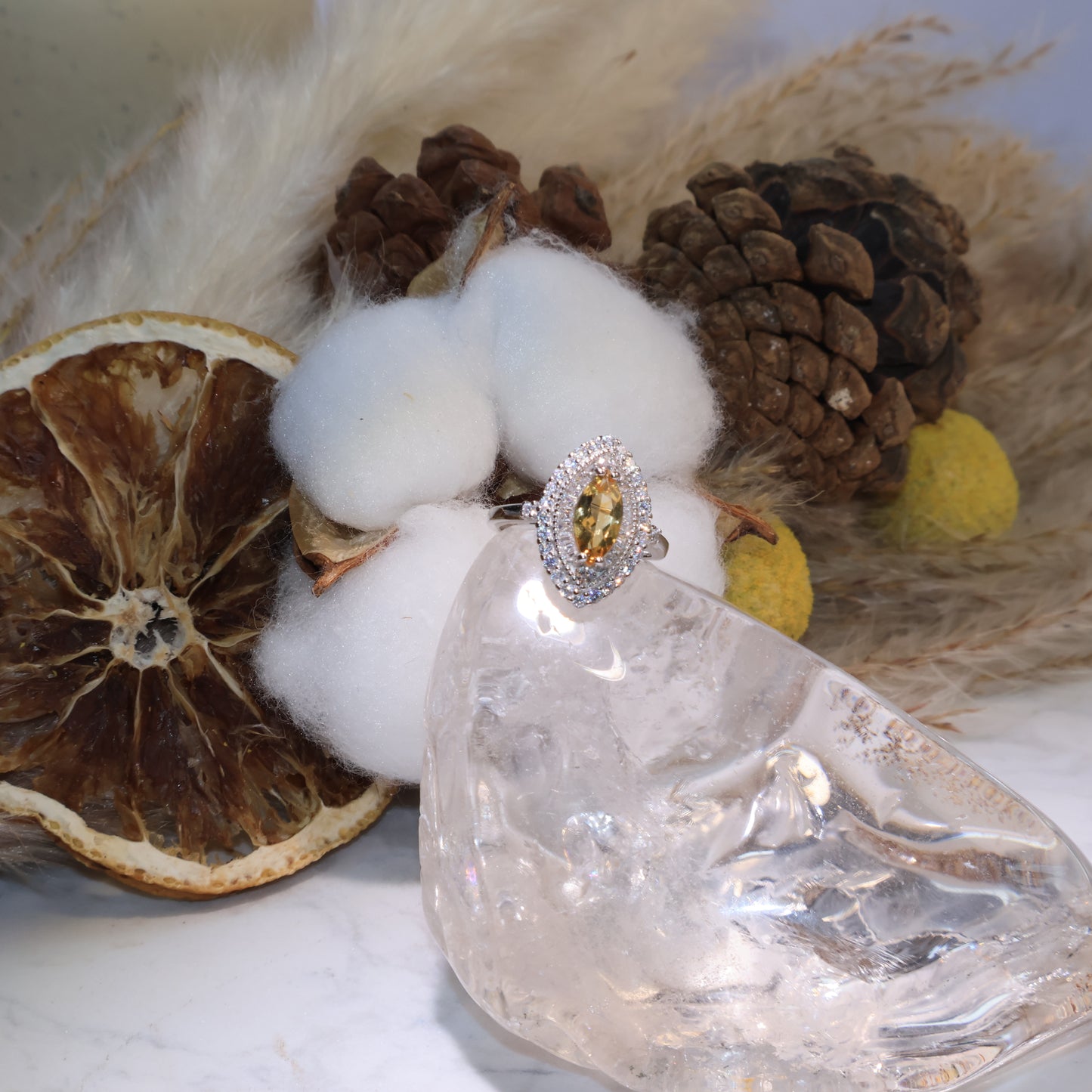 Marquise-cut citrine ring displayed on a clear quartz stand with cotton and pinecone background, glowing with golden brilliance.