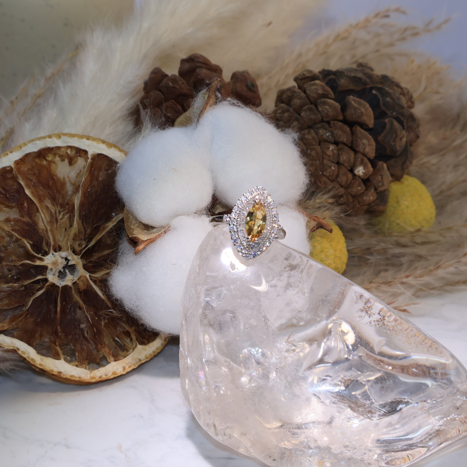 Marquise-cut citrine ring displayed on a clear quartz stand with cotton and pinecone background, glowing with golden brilliance.