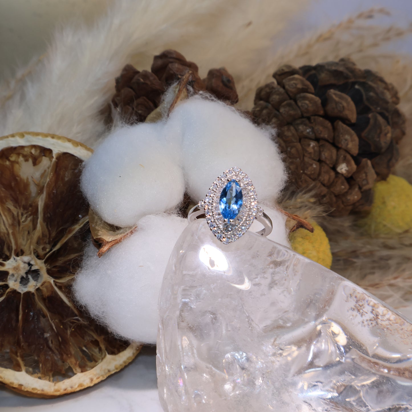 Marquise-cut blue topaz ring displayed on a clear quartz stand with cotton and pinecone background, shining with icy blue brilliance.