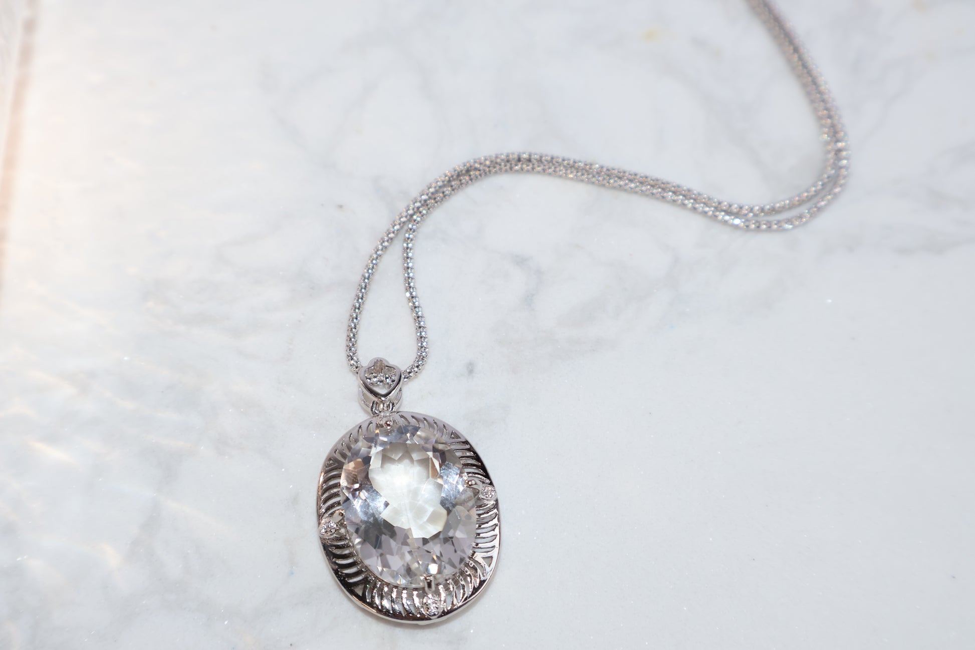 Clear quartz pendant on silver setting, oval cut, laid on marble surface
