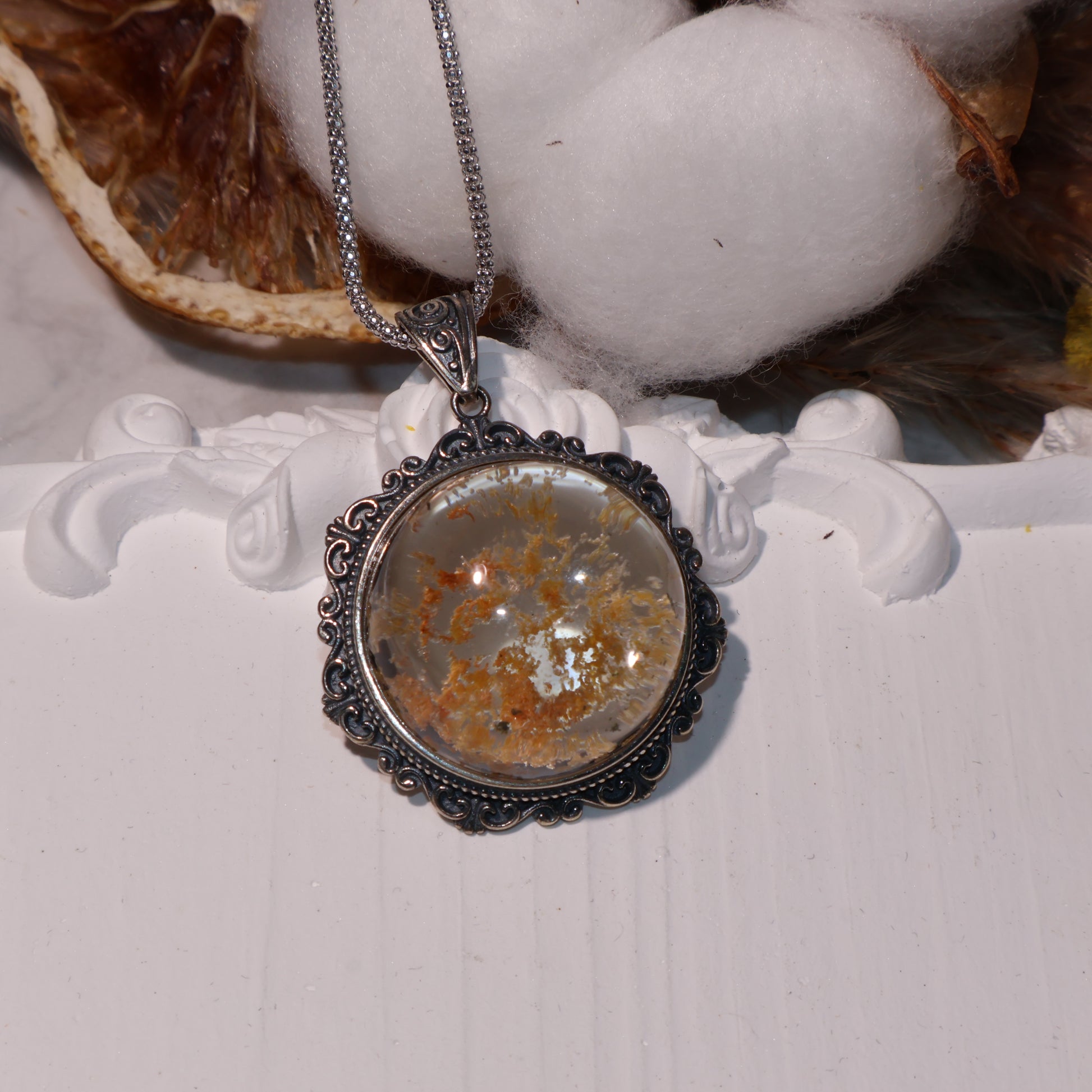 Round Phantom Quartz pendant with orange and black inclusions, set in ornate vintage-style silver frame, displayed on white floral platform.