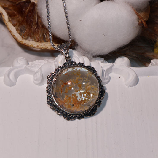 Round Phantom Quartz pendant with orange and black inclusions, set in ornate vintage-style silver frame, displayed on white floral platform.