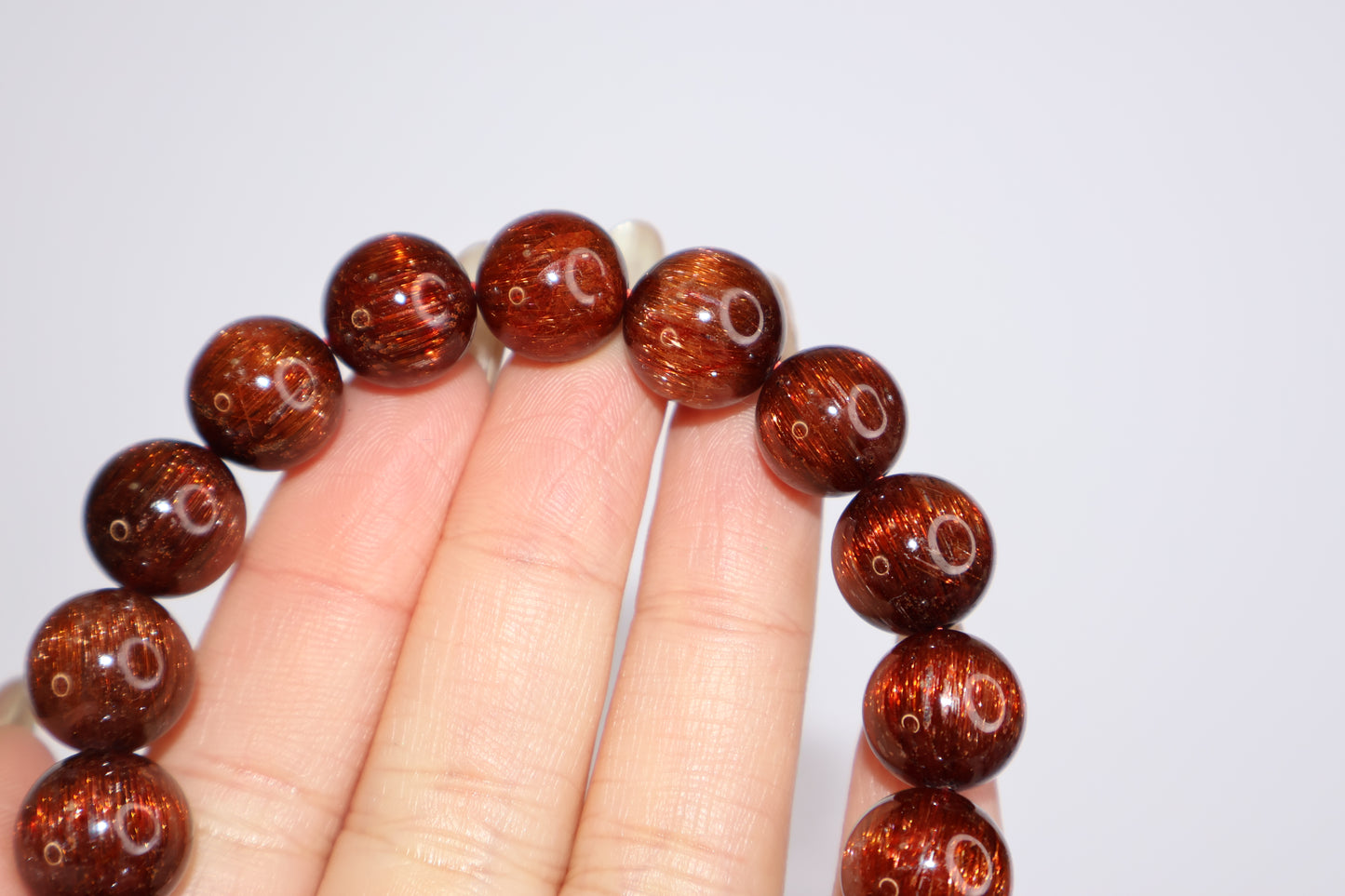 Red Rutilated Quartz beads with vivid internal rutile threads, shown between fingers to emphasize clarity, brilliance, and rich reddish-brown color with cat’s eye sheen.