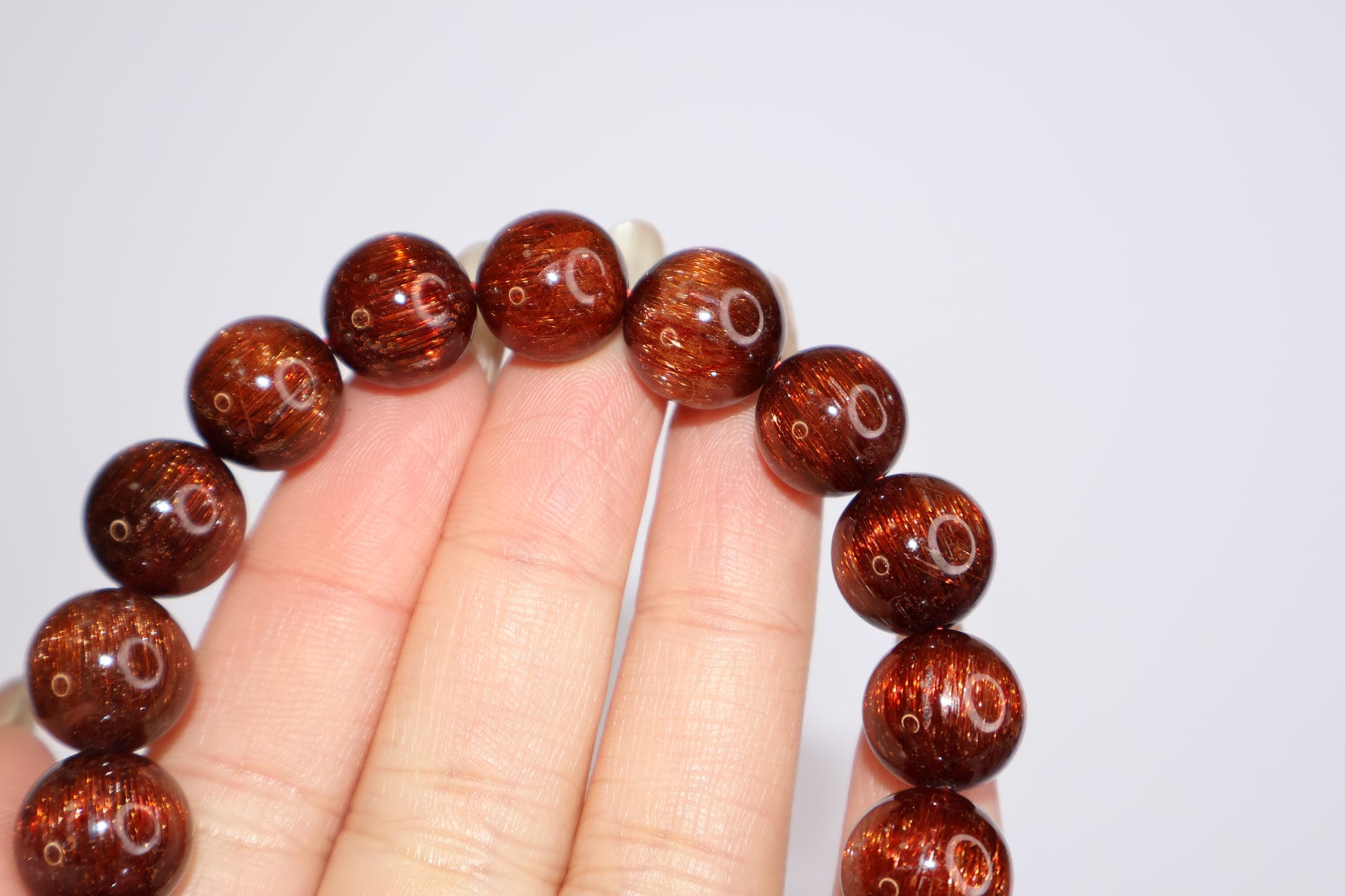 Red Rutilated Quartz beads with vivid internal rutile threads, shown between fingers to emphasize clarity, brilliance, and rich reddish-brown color with cat’s eye sheen.